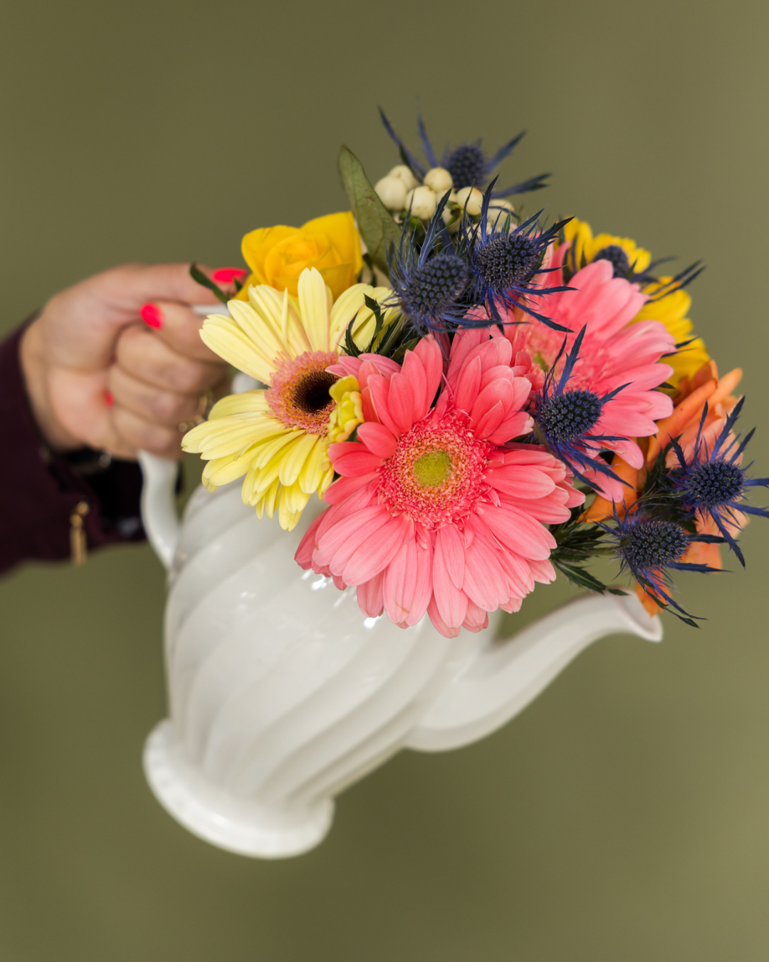 Person holding a white ceramic vase with a colorful bouquet of pink, yellow, and orange flowers and blue thistles, against a green background.
