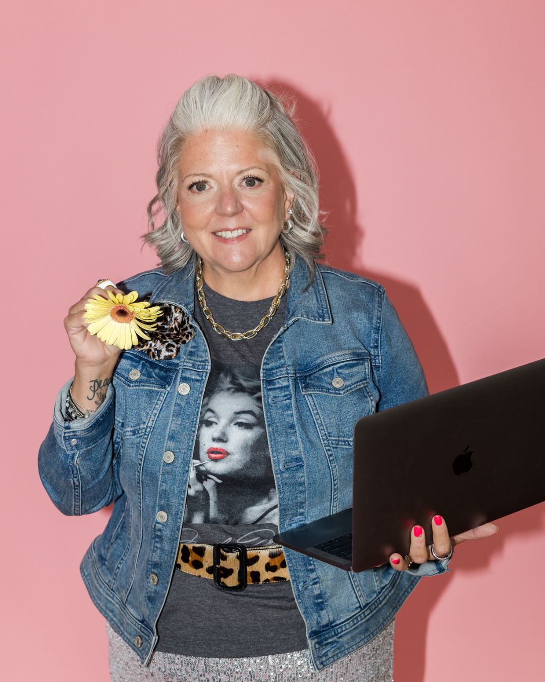 A woman with gray hair styled in waves, wearing a denim jacket over a Marilyn Monroe graphic T-shirt, holding a yellow daisy flower and leopard print scrunchie, smiling, standing against a pink background with a laptop in her hand.