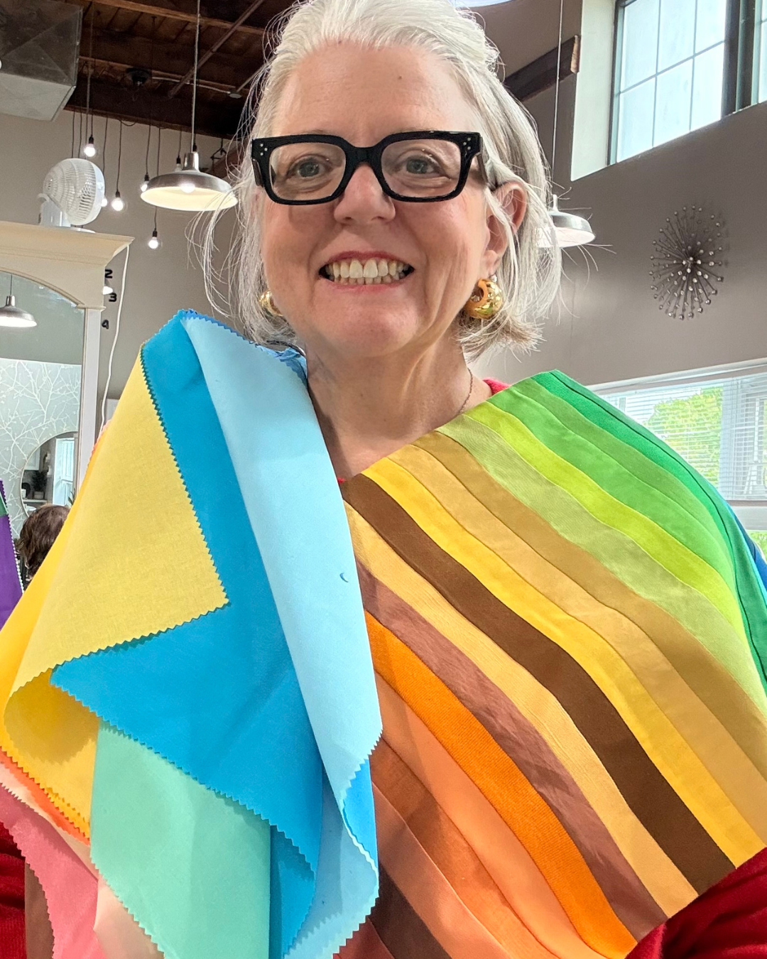 Woman with gray hair, black glasses, and gold earrings smiling, wearing a rainbow-colored sash, holding rainbow-colored cloths, inside a well-lit room with industrial-style lighting.