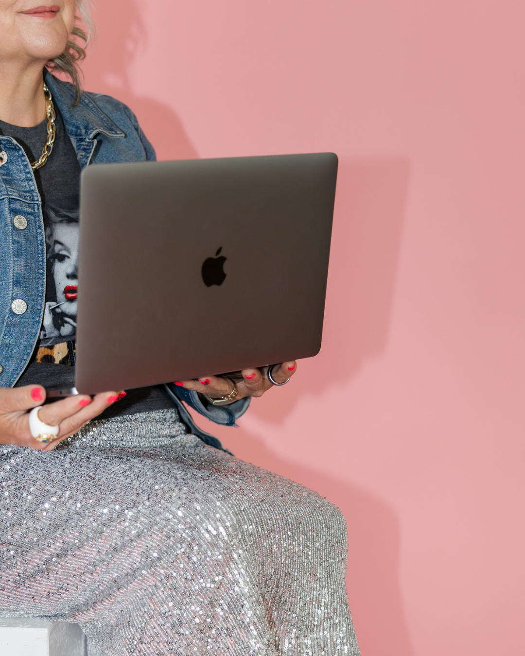 A woman sitting against a pink background, holding a silver MacBook laptop.