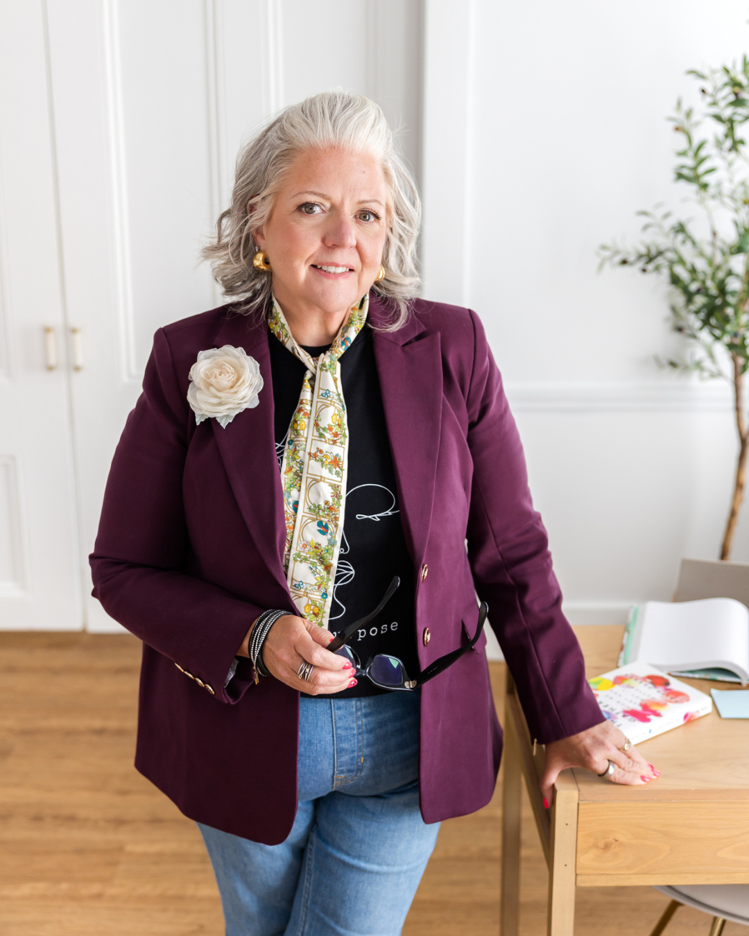 A smiling woman with gray hair dressed in a purple blazer, holding glasses, standing in a bright room with a table and books in the background.