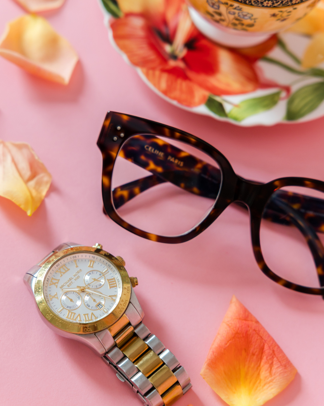 A Michael Kors wristwatch with a silver and gold band, a round white face with Roman numerals, and multiple smaller dials, a pair of tortoiseshell glasses, a colorful floral teacup and saucer in the background, and rose petals on a pink surface.