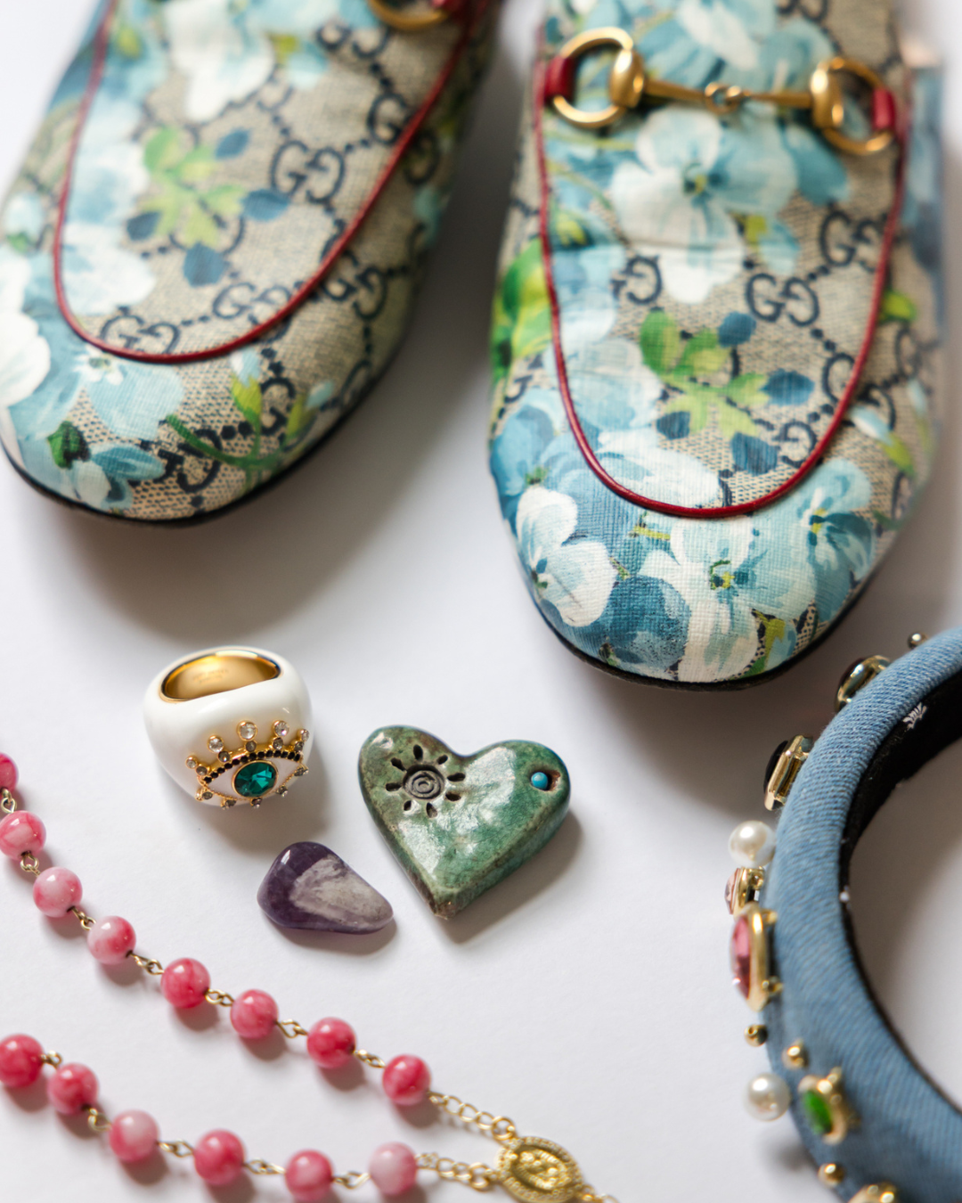 Pair of floral-patterned slip-on shoes with gold and red accents, decorated with Gucci logo, along with jewelry and decorative objects such as a pink beaded necklace, a pearl bracelet, a heart-shaped ceramic piece, a small purple stone, a gold ring with an eye design, and a blue and black fabric bracelet with pearl embellishments.