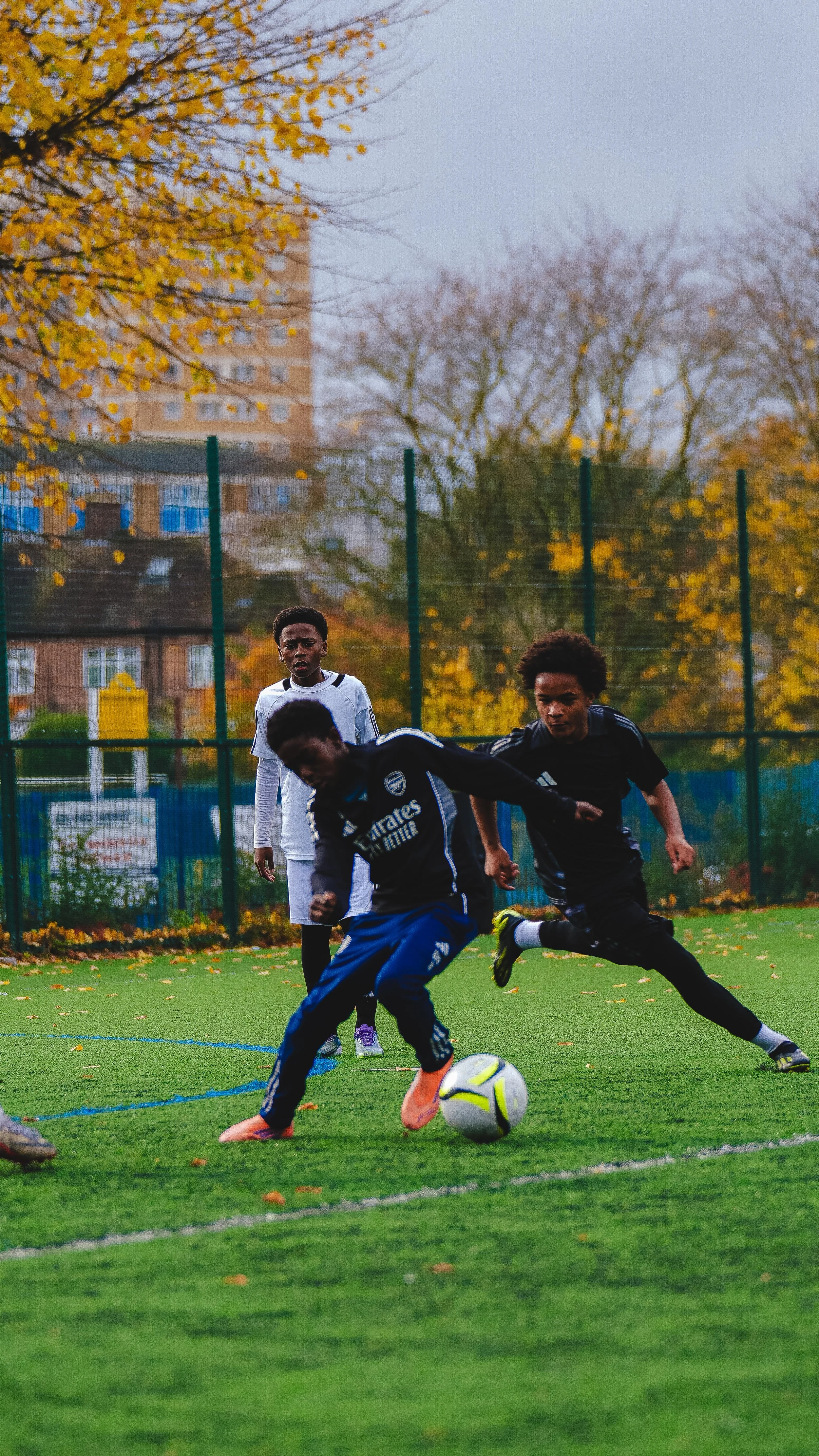 Three boys playing football on a pitch, with fall trees and a building in the background.