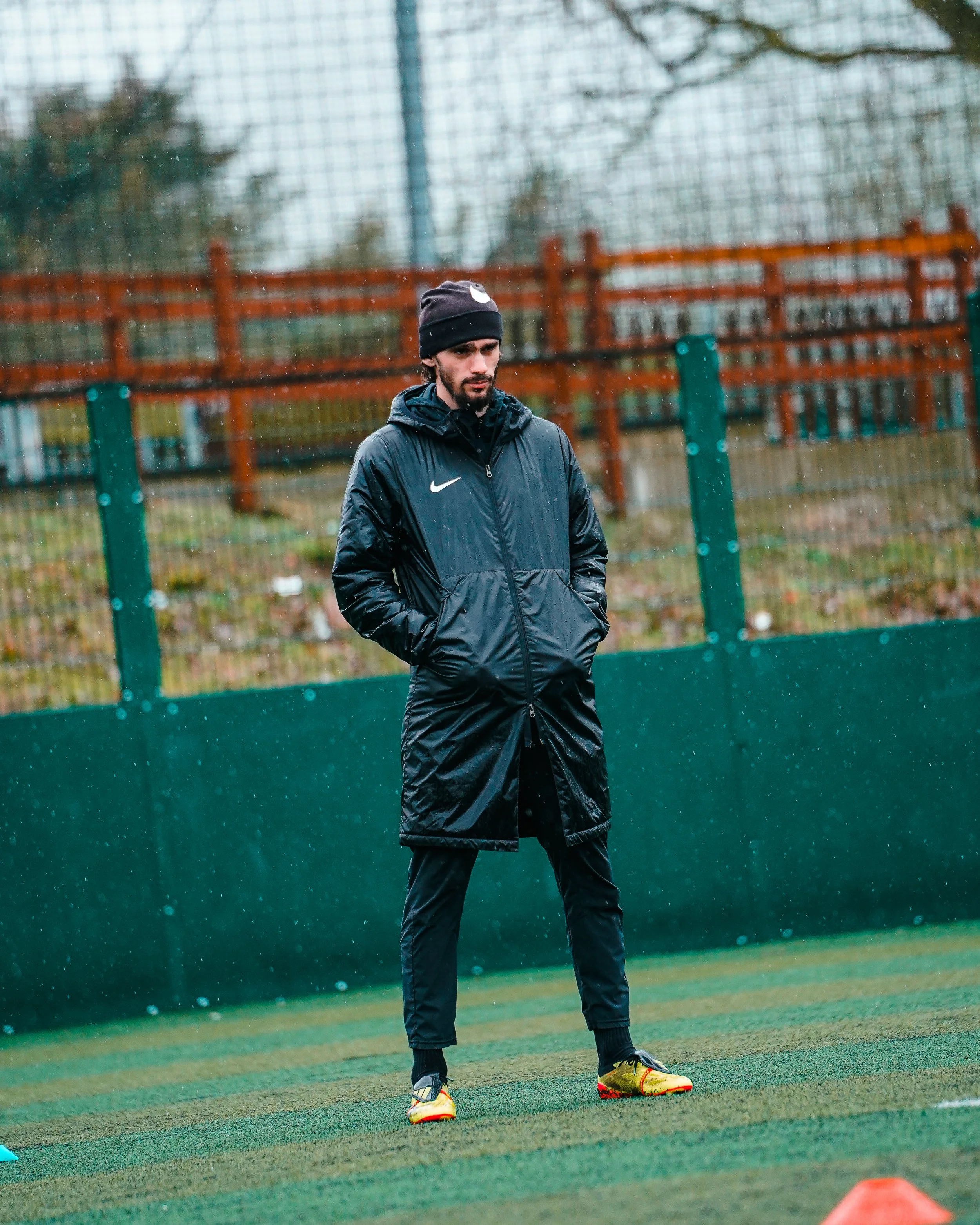Man standing on a football pitch in rainy weather, wearing black sportswear, a black beanie, and cleats, with a fencing or netted fence in the background.
