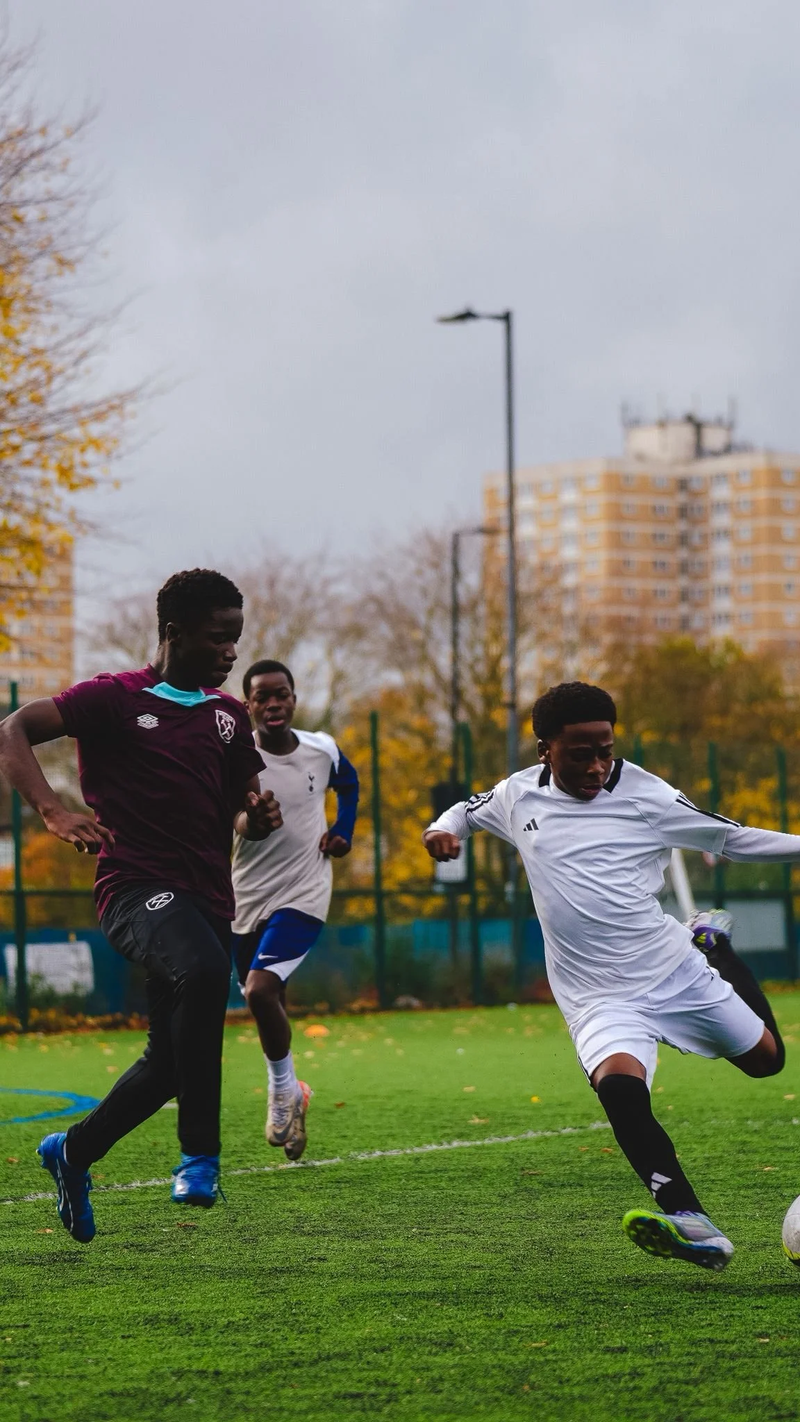 Three young boys playing soccer on a green field during overcast weather with trees and a tall building in the background.