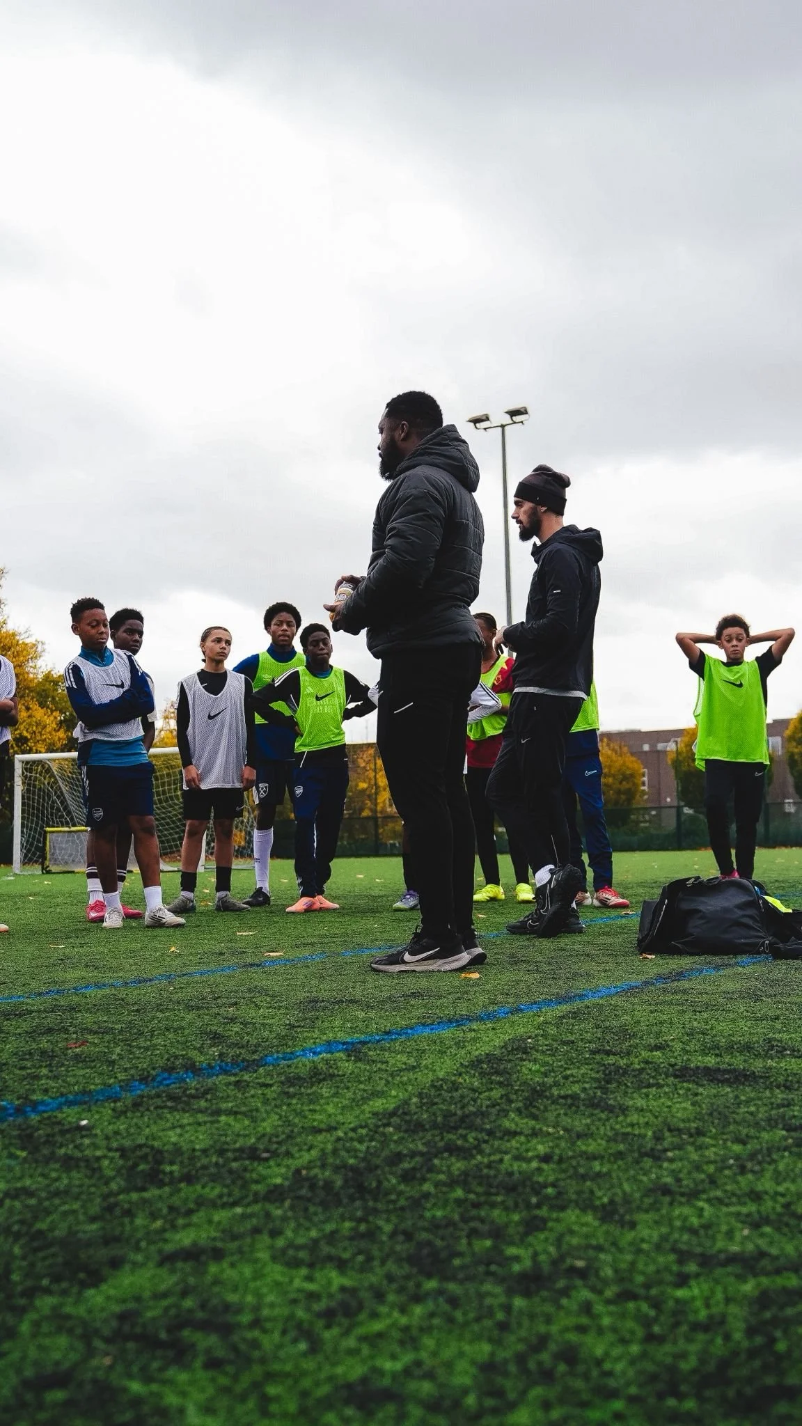 Football coach giving instructions to young players during practice on an outdoor football field, with players wearing training bibs and listening attentively.