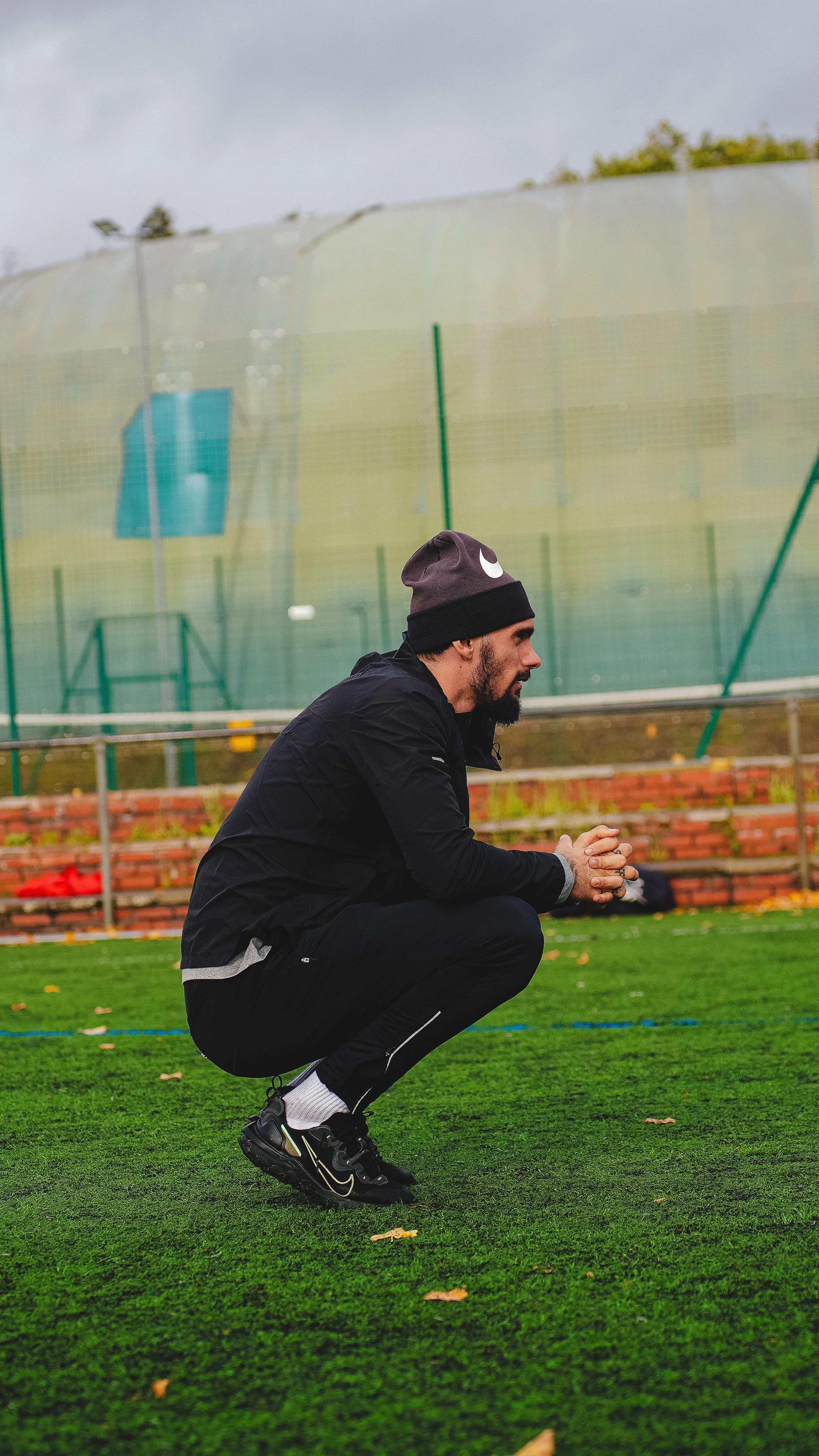 Man squatting on grass field outdoors in rainy weather, wearing black jacket, black pants, black sports shoes, gray socks, and gray beanie with black brim, with a large green structure in the background.