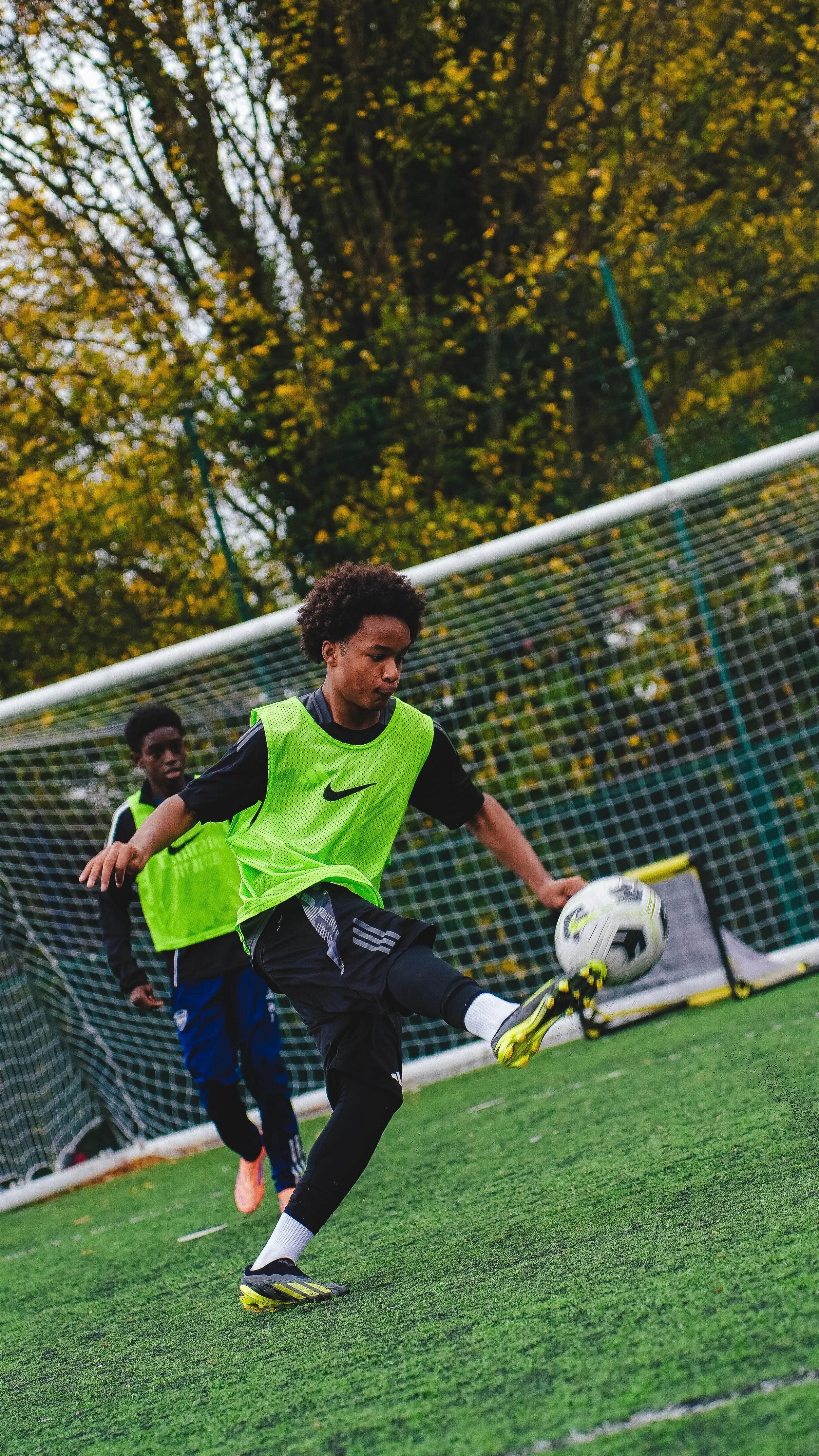 Two young boys playing soccer on a field, one kicking a soccer ball, during daytime with trees in the background.