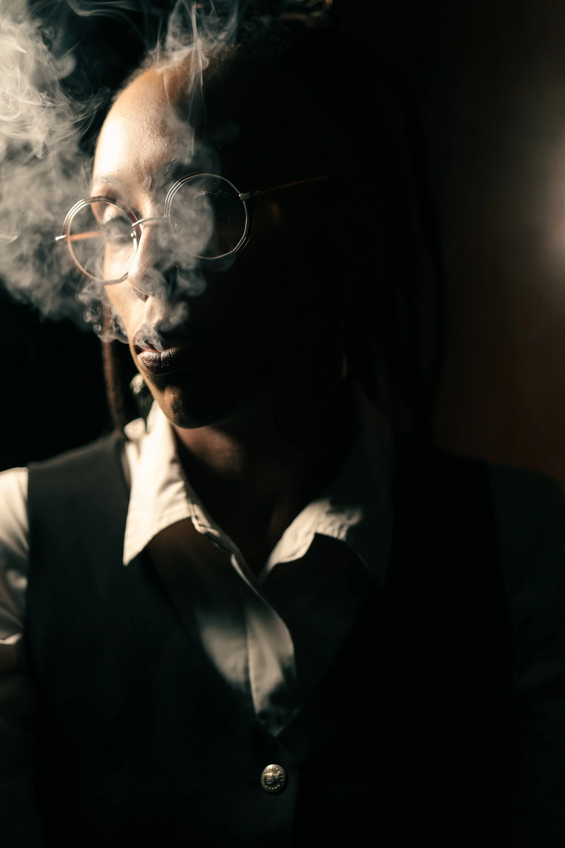 A person with glasses and a black vest over a white collared shirt, smoking with smoke visible in front of their face, dark background.