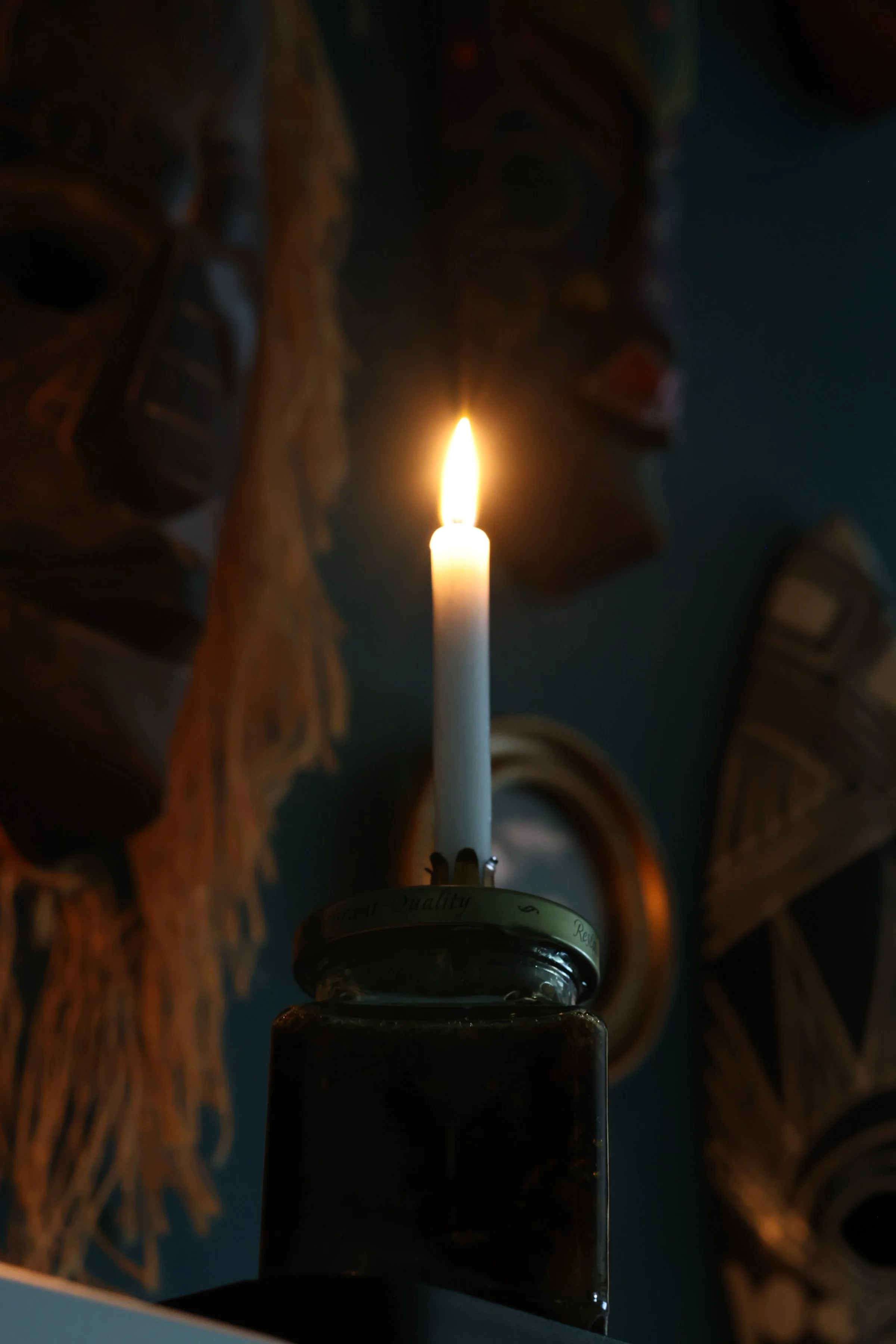 A lit candle held in a dark room. The background features large, carved wooden letters spelling out words, possibly "STOP" or similar, illuminated softly by the candle's glow.