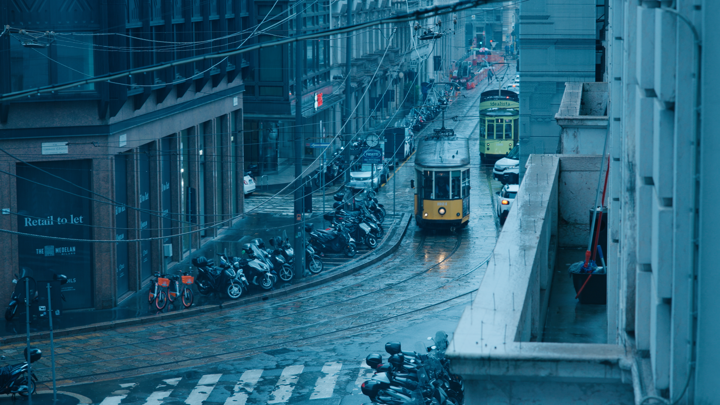 Cinematic overhead shot of a rain-soaked city street with trams, a location filming still from an atmospheric book trailer produced for a UK publisher by Gunpowder House Productions