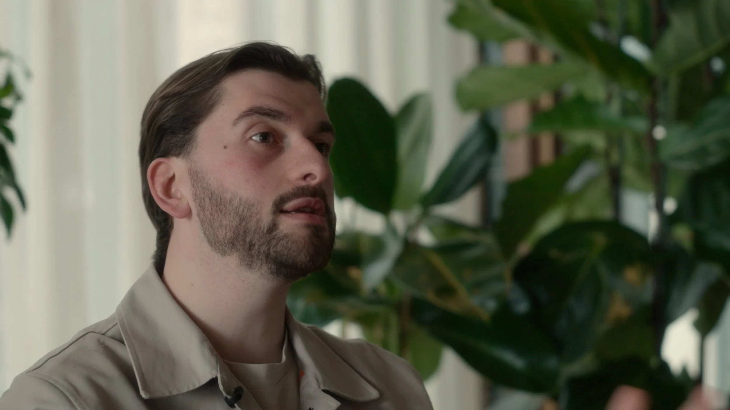 A man with dark hair and a beard, wearing a beige shirt, standing near large green houseplants.