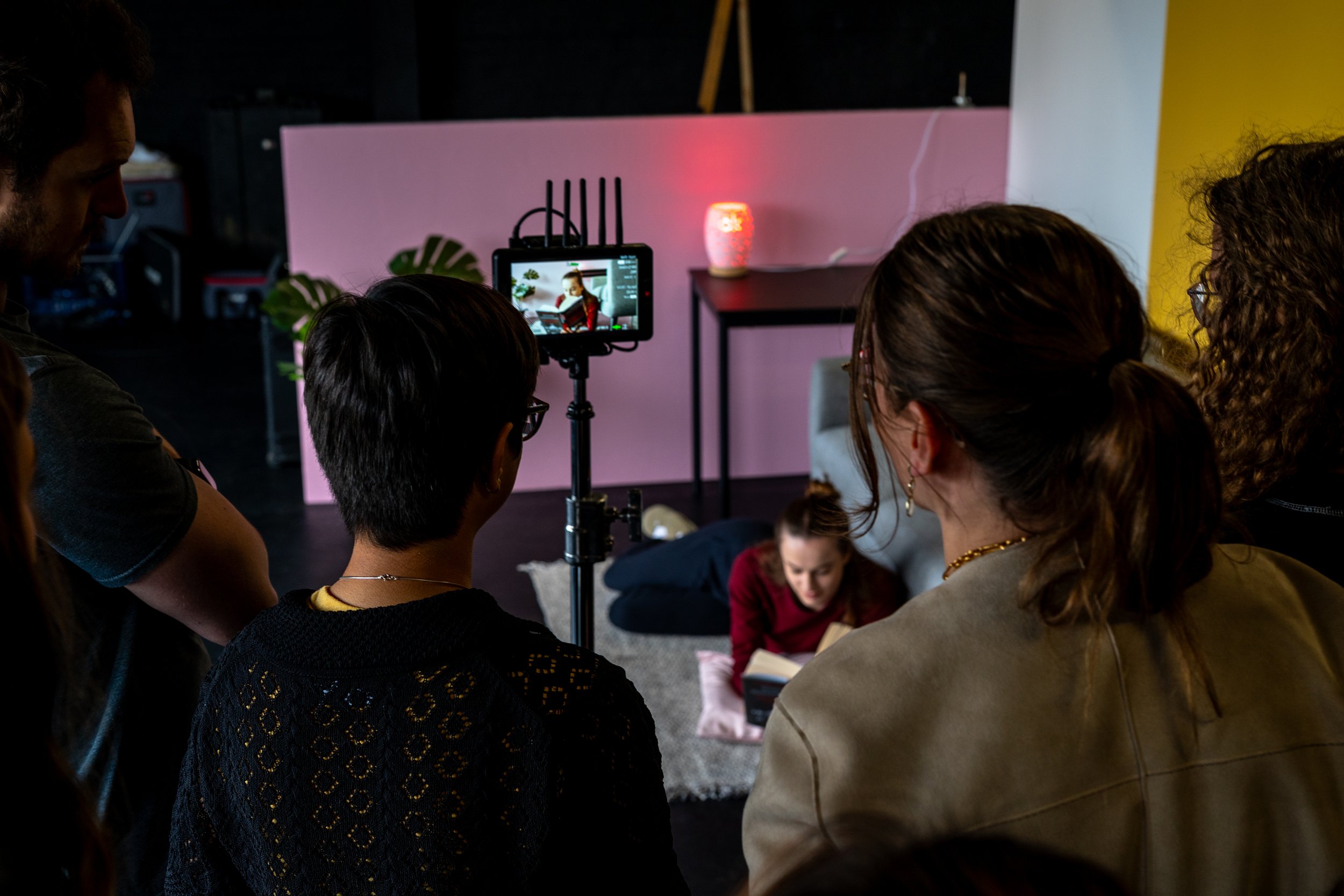 Group of people gathered around a camera on a tripod, filming or photographing a woman reading a book on the floor in a cozy indoor setting with pink and yellow walls and decorative lighting.