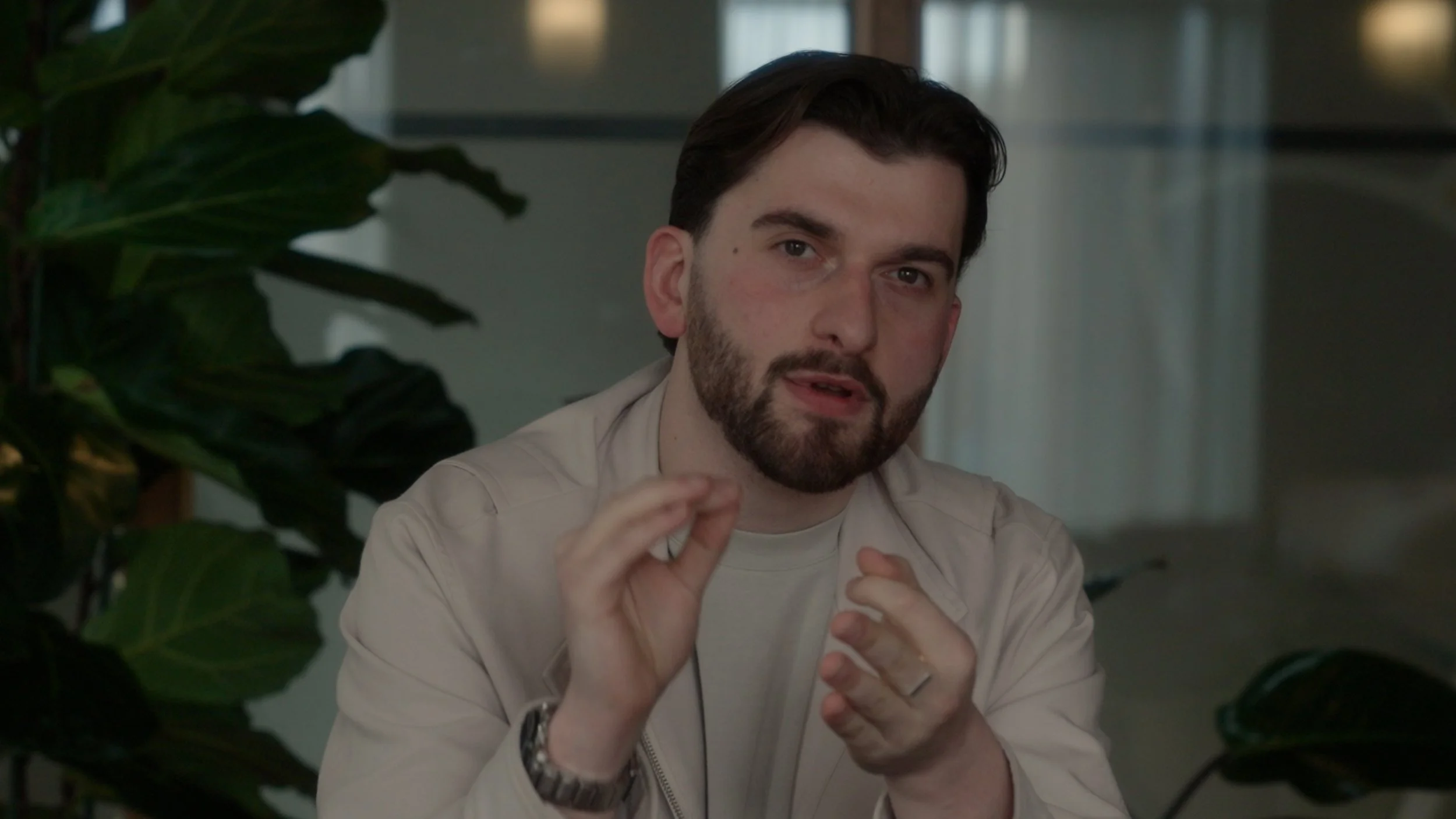 A man with dark hair and a beard, wearing a beige jacket and white shirt, is speaking and gesturing with his hands, seated indoors with a large green plant on the left.