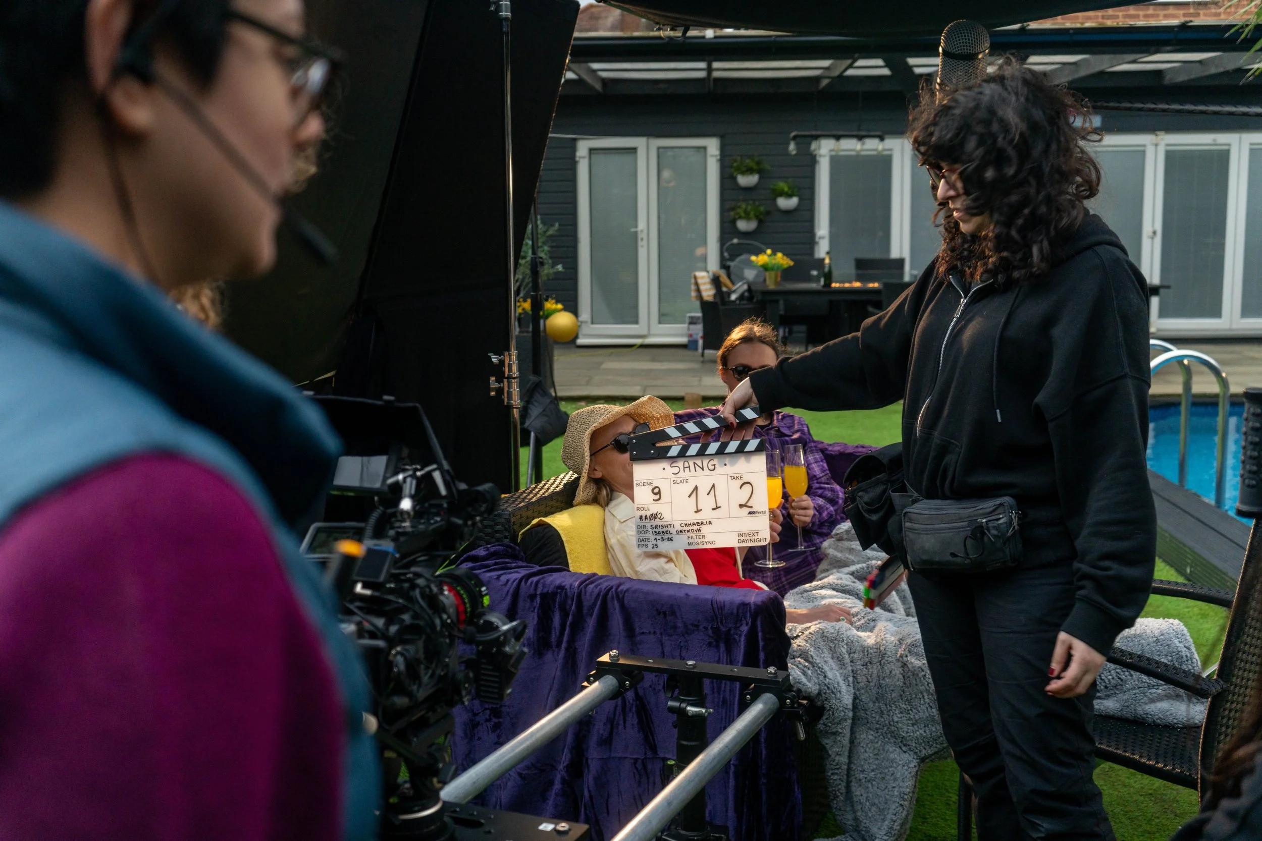 A woman with curly dark hair and glasses is adjusting a clapperboard in front of a woman dressed as an elderly person lying on a lounge chair, wearing sunglasses, a straw hat, and a yellow scarf. The scene is set outdoors with a house and garden in the background during what appears to be a film or television shoot.