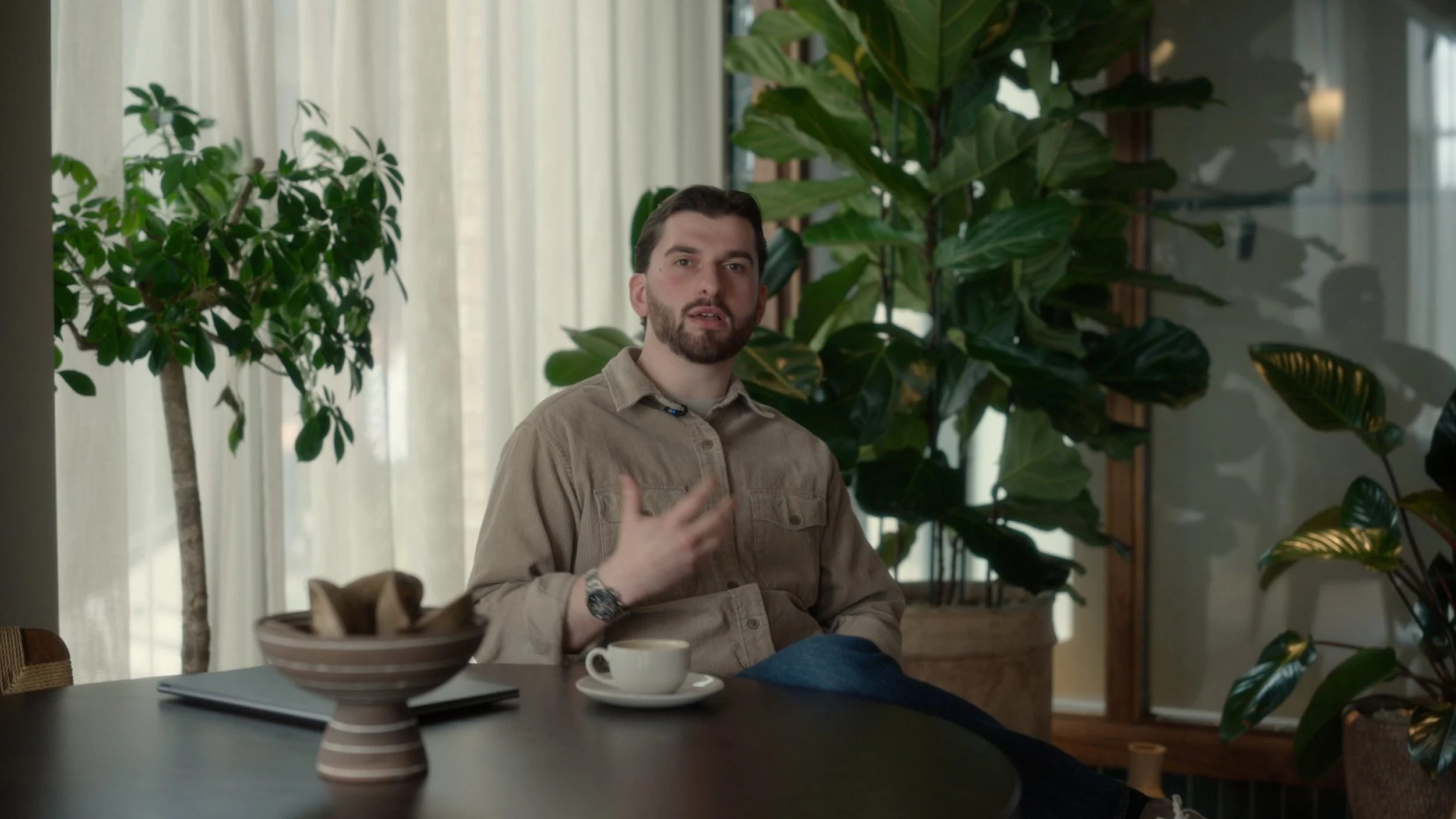 A man with a beard and brown hair is sitting at a table in front of large green plants. He is wearing a beige shirt and a watch, and he appears to be speaking. On the table, there is a bowl and a cup. Light filters through sheer curtains in the background.