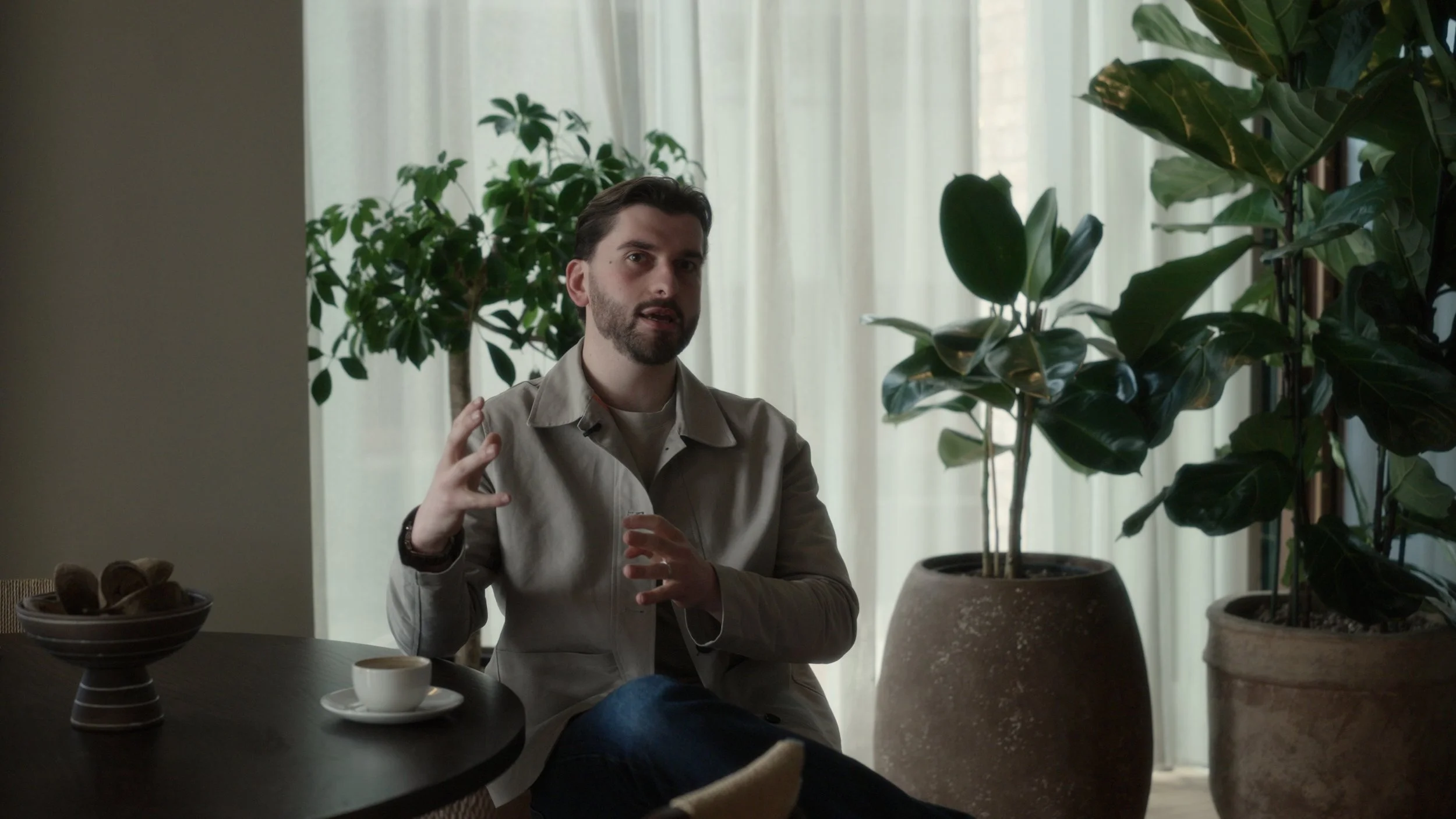 A man with dark hair, beard, and wearing a light-colored shirt, sitting in a room with large green potted plants, a curtain in the background, and a table with a cup and a bowl in front of him.