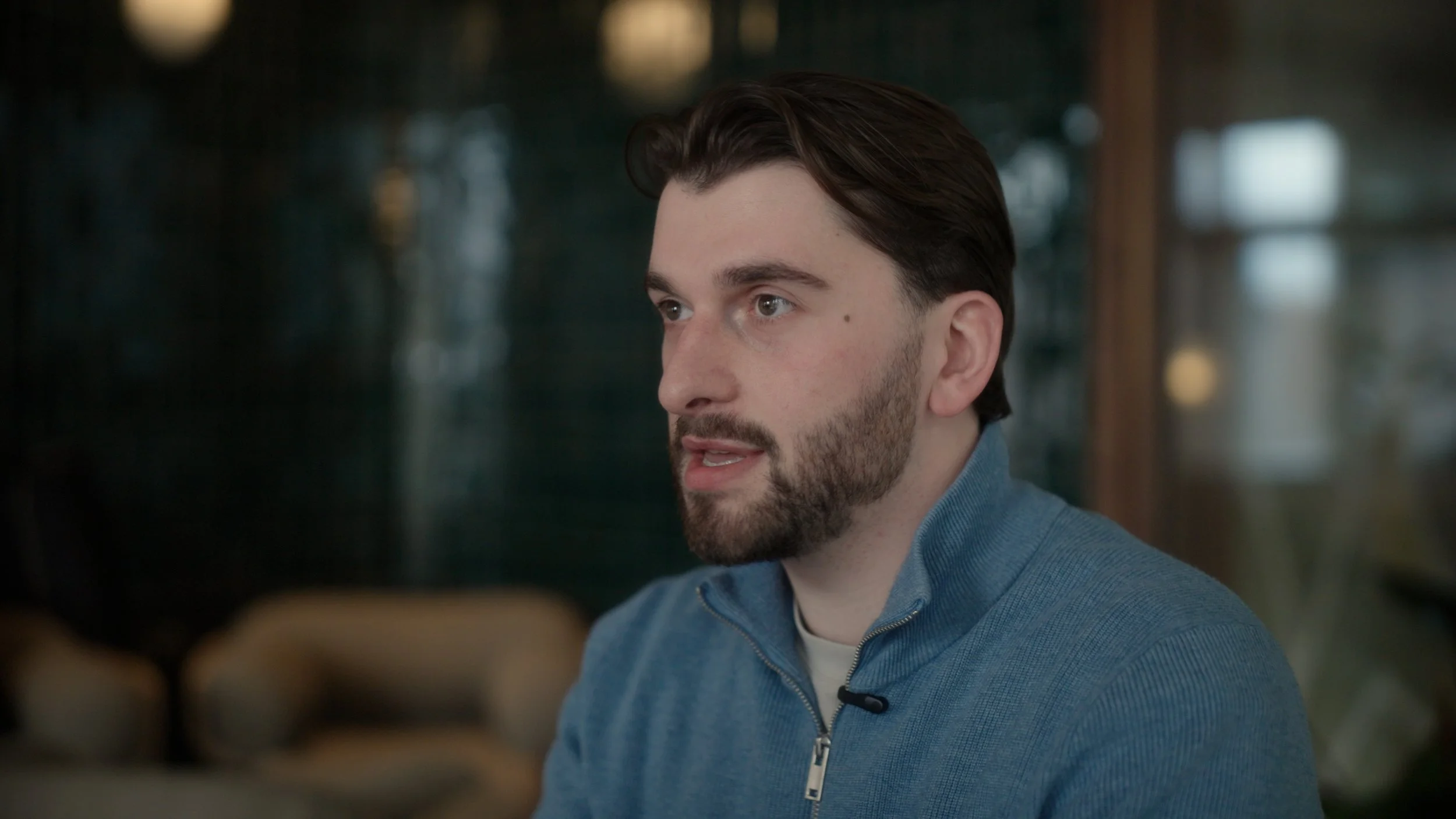 A young man with dark hair and a beard, wearing a blue zip-up sweater, is seated indoors and appears to be engaged in conversation.