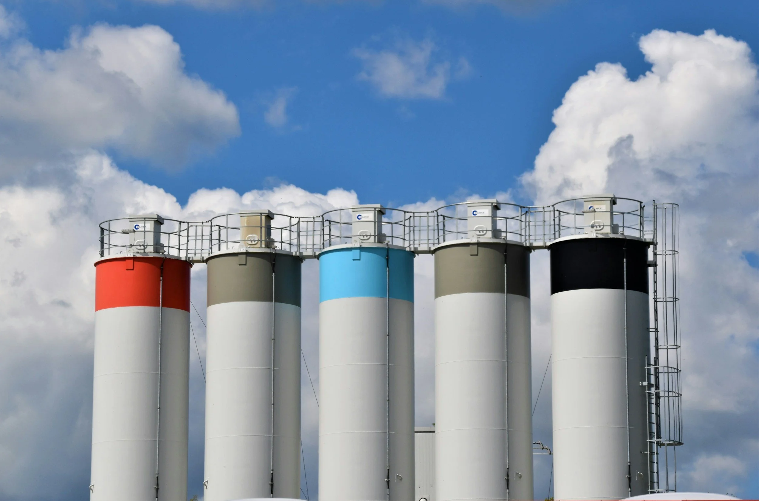 Five industrial silos with colored tops in red, beige, blue, beige, and black against a cloudy sky.