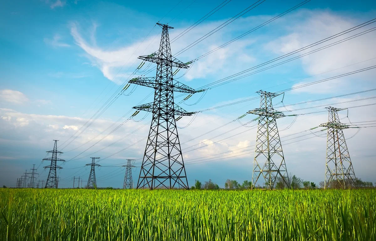A field of green grass with multiple power transmission towers and lines under a partly cloudy blue sky.