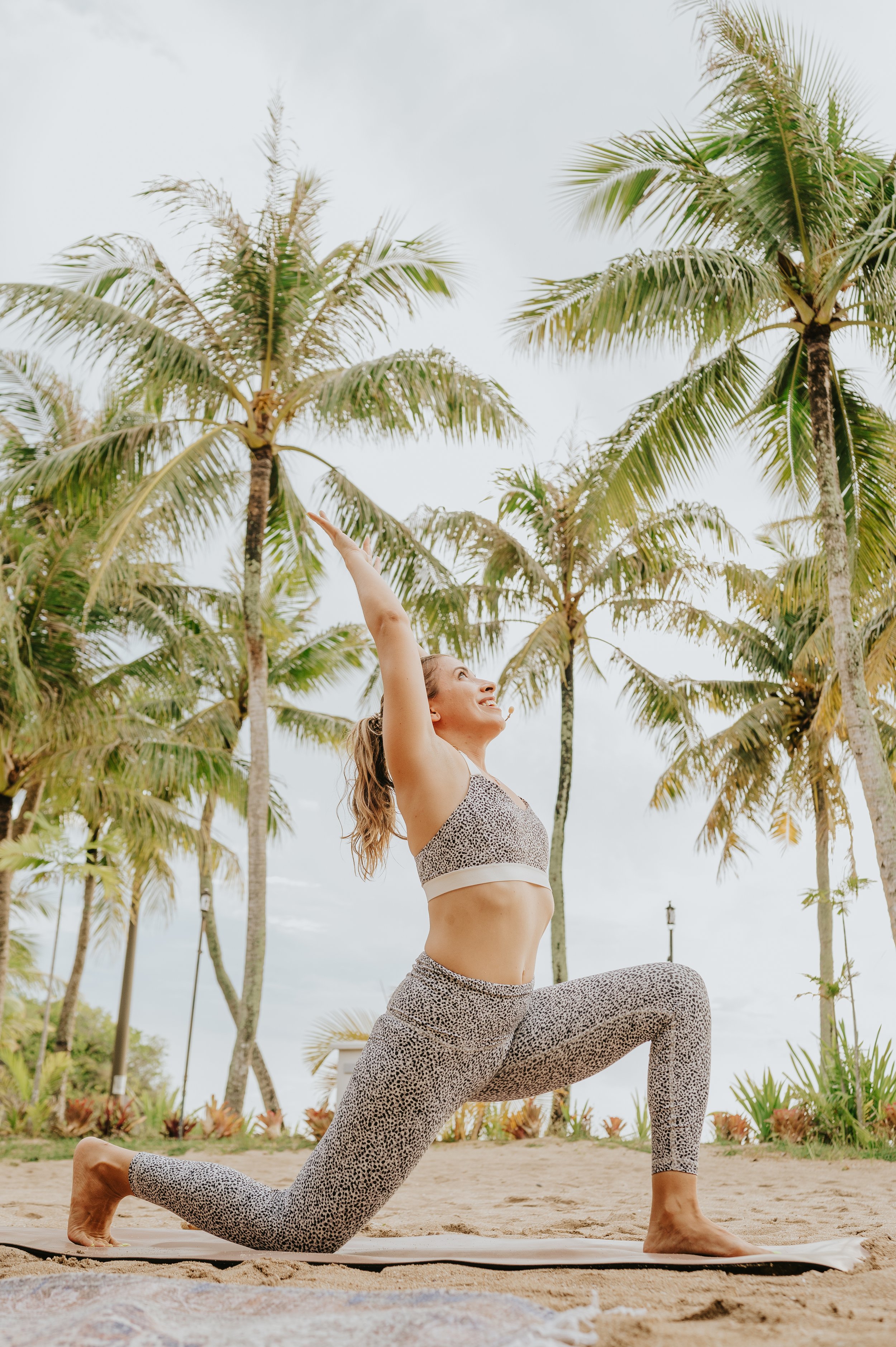 A woman practicing yoga outdoors on a sandy beach, surrounded by tall palm trees, performing a low lunge pose with arms extended upward, wearing a matching leopard print sports bra and leggings.