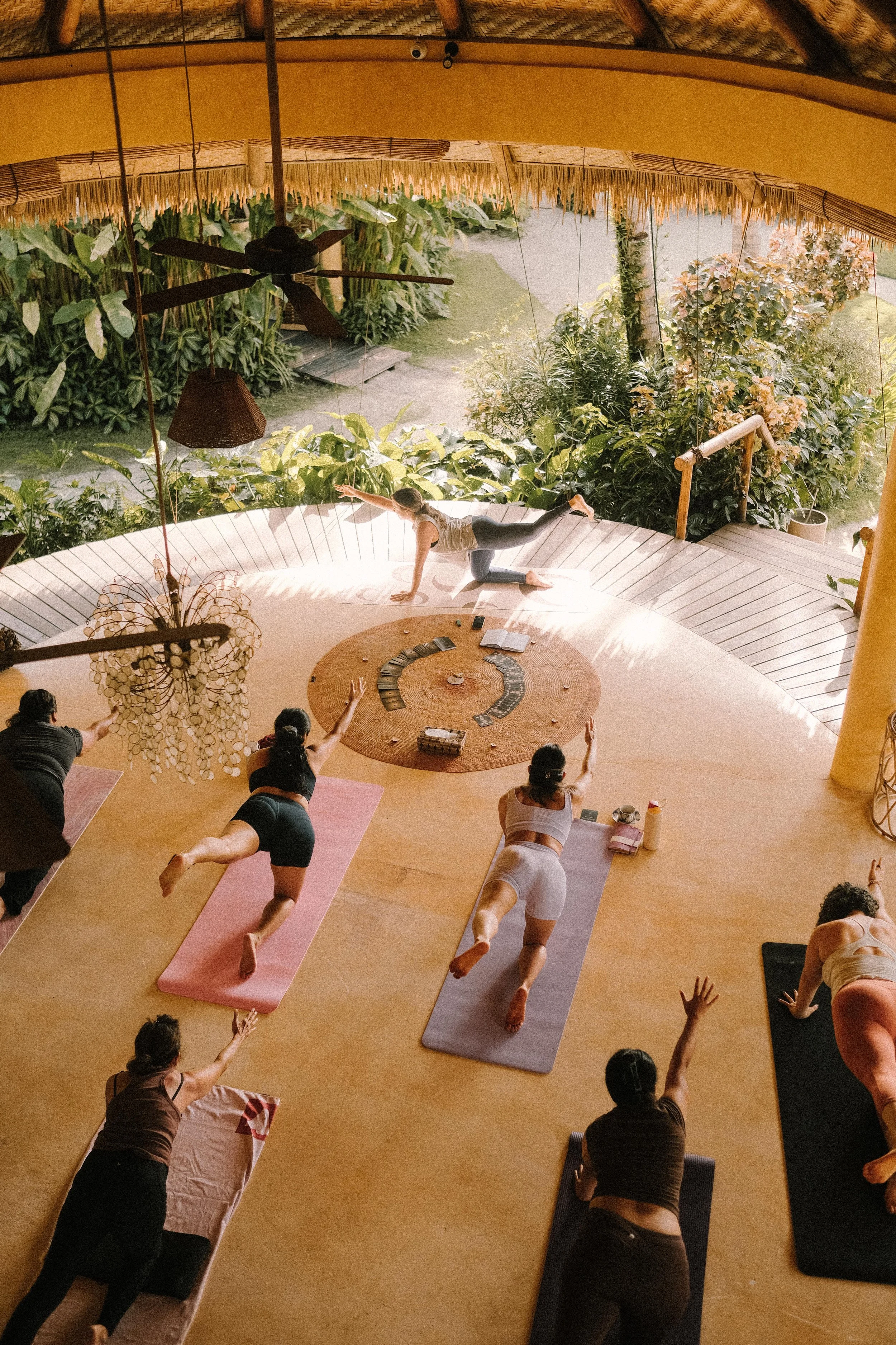 People participating in an outdoor yoga class on a deck, standing with arms raised, facing a scenic green landscape and cloudy sky.