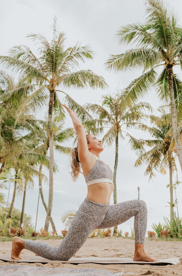 A woman practicing yoga outdoors on a sandy beach, surrounded by tall palm trees, performing a low lunge pose with arms extended upward, wearing a matching leopard print sports bra and leggings.