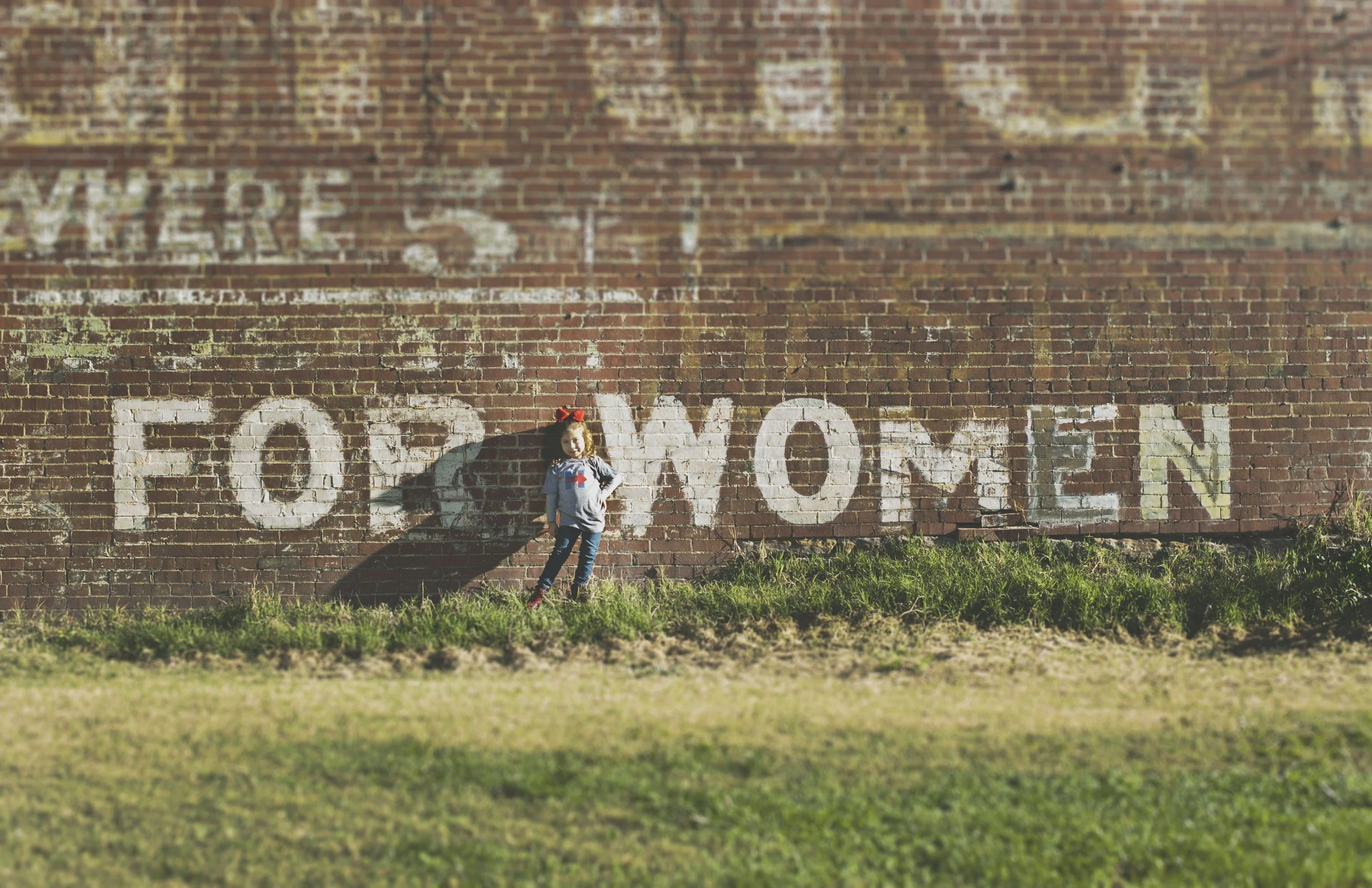 Ein Mädchen steht vor einer alten Brickwand mit dem Wort 'FREIWILLIG' in großen weißen Buchstaben. Das Mädchen trägt ein graues T-Shirt mit einem roten und blauen Design, blaue Jeans und rote Schuhe, und hat eine rote Schleife im Haar.