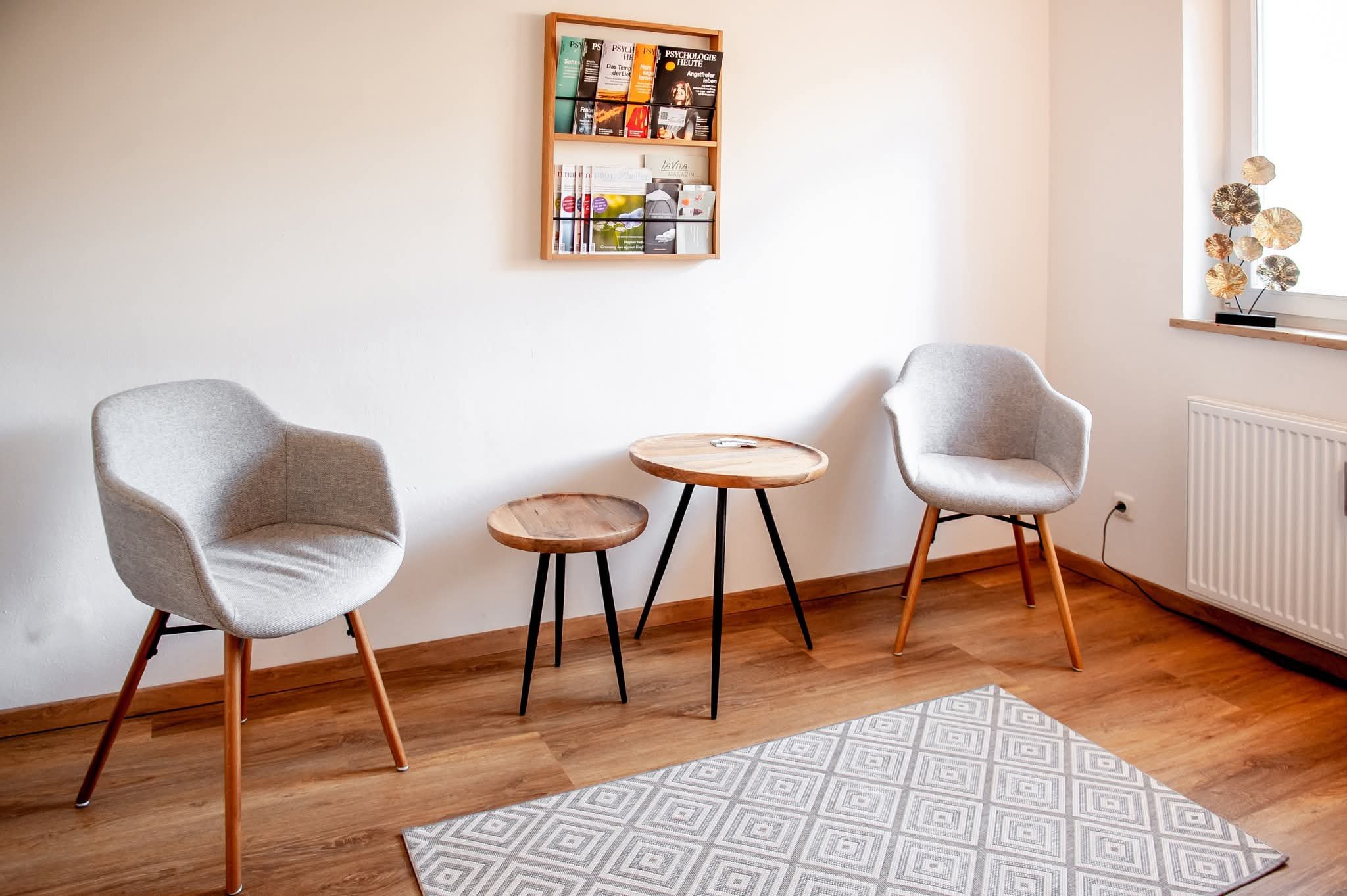 Zwei moderne, graue Sessel mit Holzbeinen, zwei kleine Holztische mit schwarzen Beinen, eine Bücherwand an der weißen Wand, ein Fenster mit Dekoration, Teppich mit geometrischem Muster auf Holzboden.