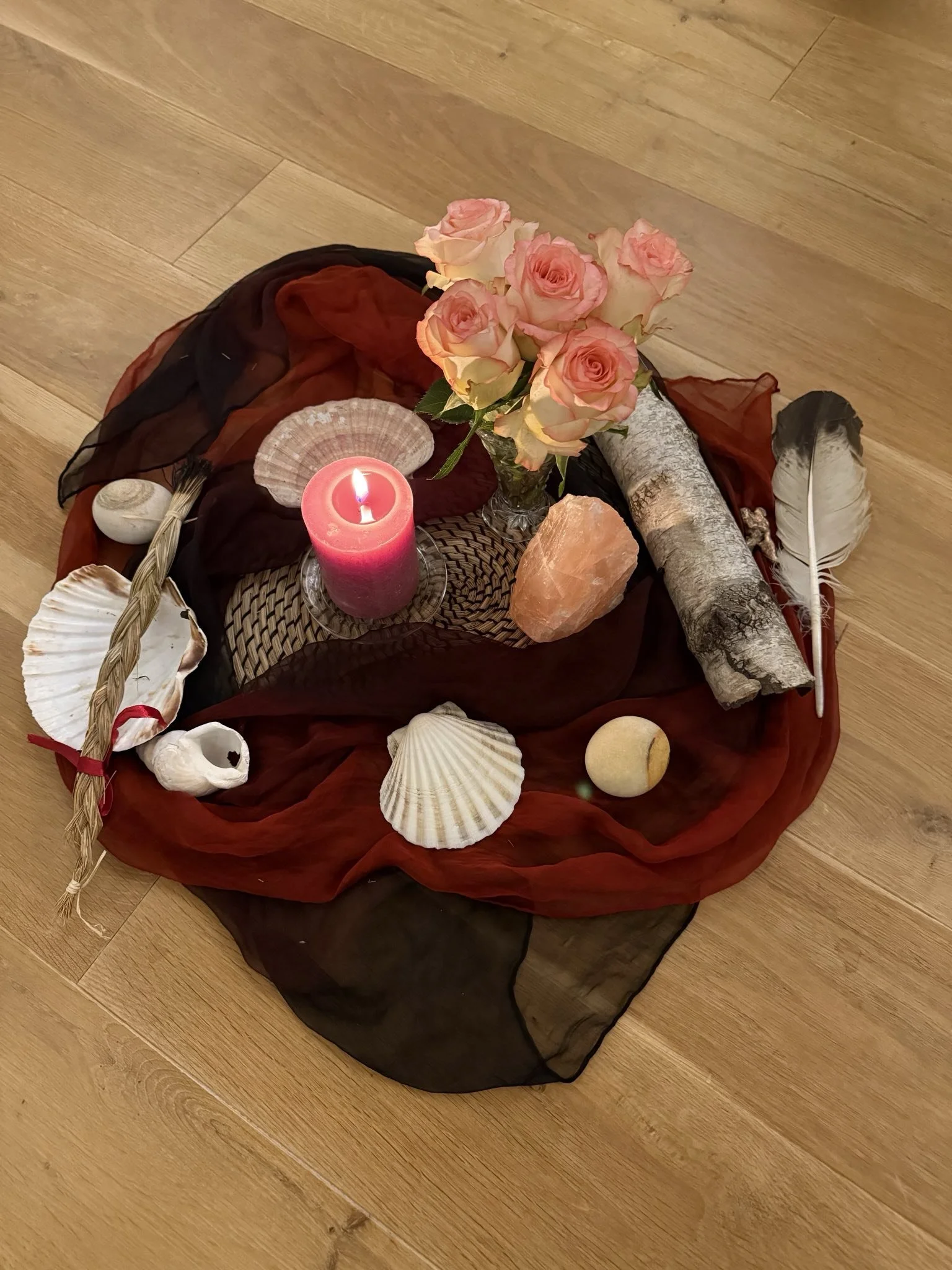 Eine Dekoration mit Rosen, Muscheln, Steinen, Federn, einer Kerze und einem Stück Holz auf einem roten und schwarzen Tuch auf Holzparkett.