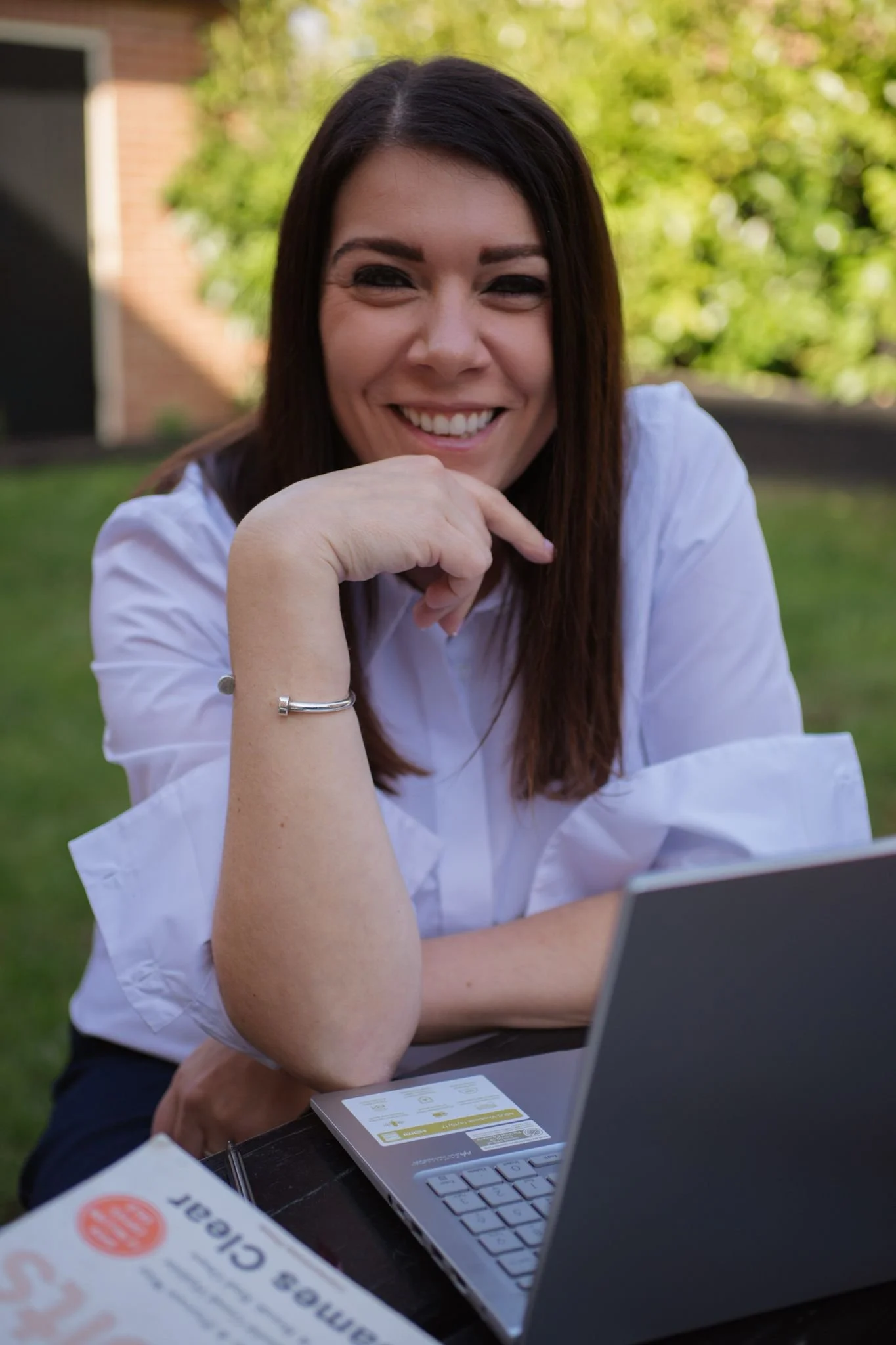 A woman with long dark hair smiling and sitting outdoors in front of a laptop, wearing a white shirt, with a newspaper on the table.