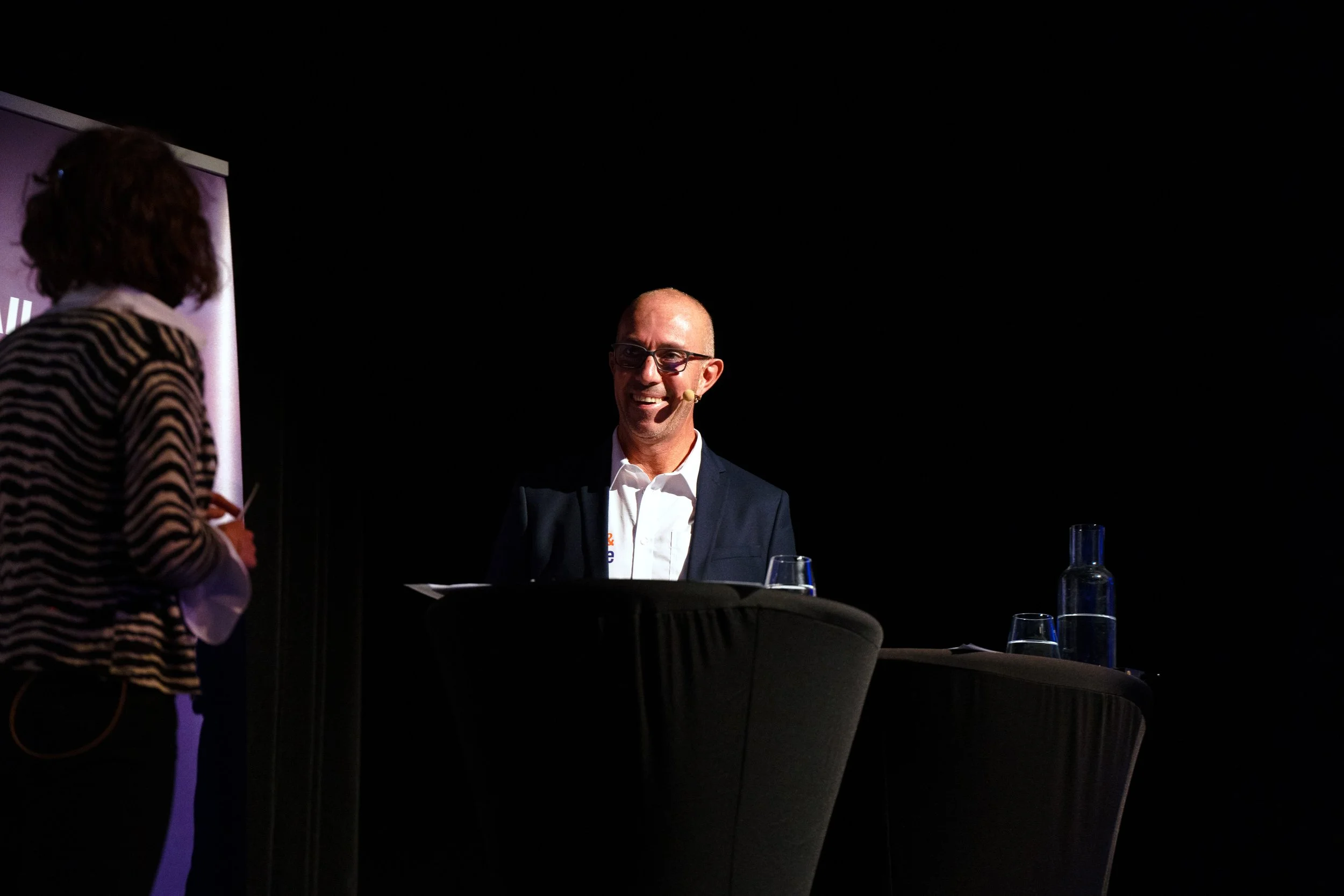 A conference scene with a smiling man, wearing glasses and a suit, seated behind a table, opposite a woman whose back and part of her silhouette are visible, in a darkened room.