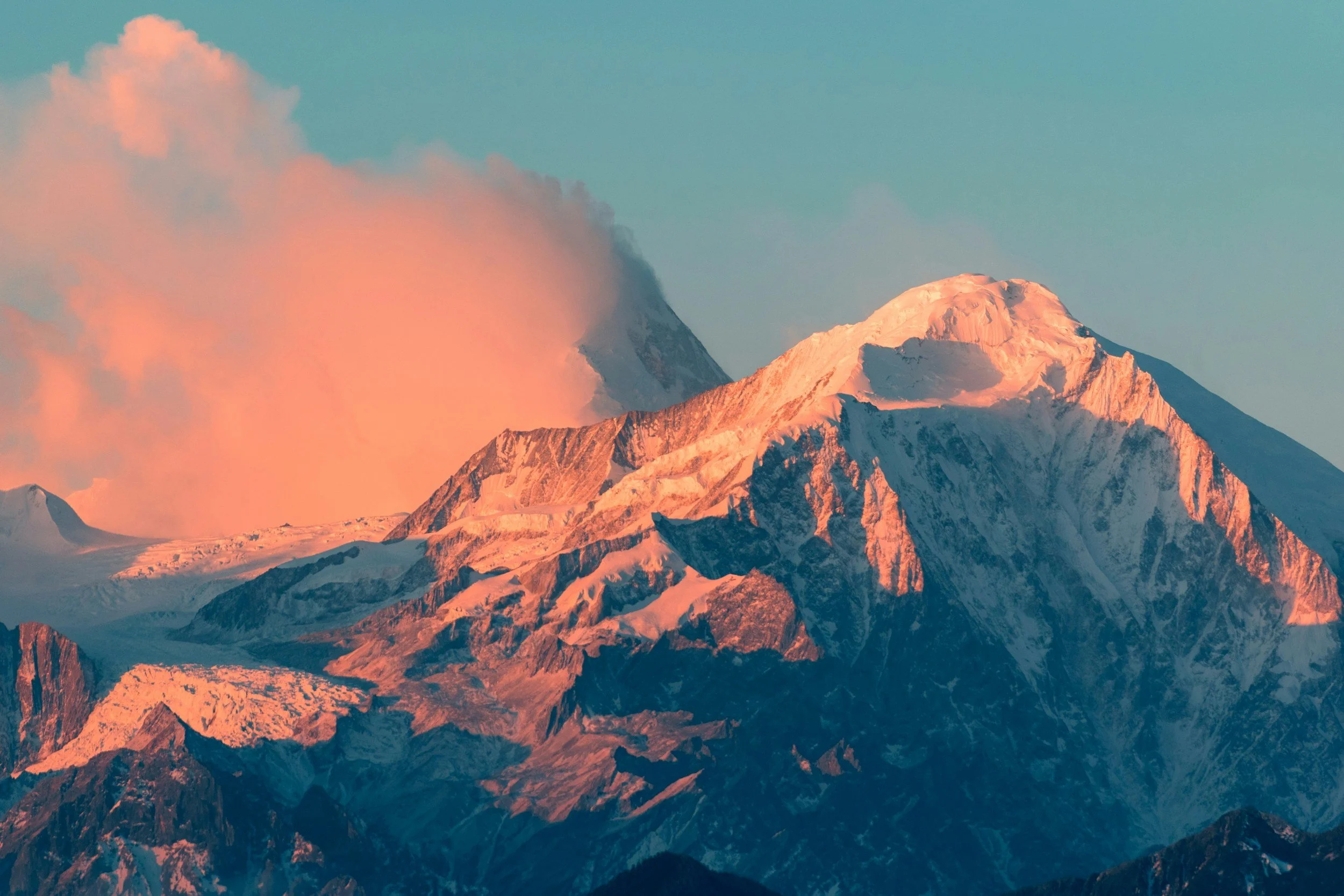 Snow-capped mountains at sunset or sunrise, with an active volcanic peak emitting smoke.