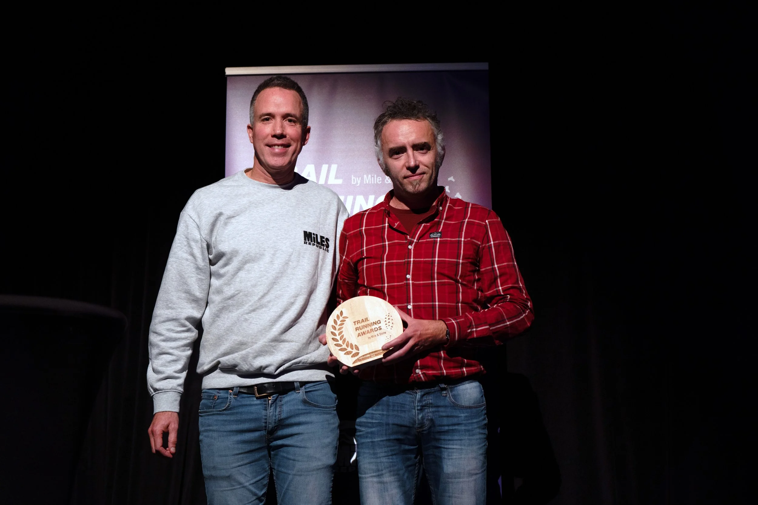 Two men stand on stage, one wearing a grey sweatshirt with the 'Miles' logo and the other a red-checkered shirt. The man in the shirt is holding a wooden trophy inscribed 'Trail Running Awards'.