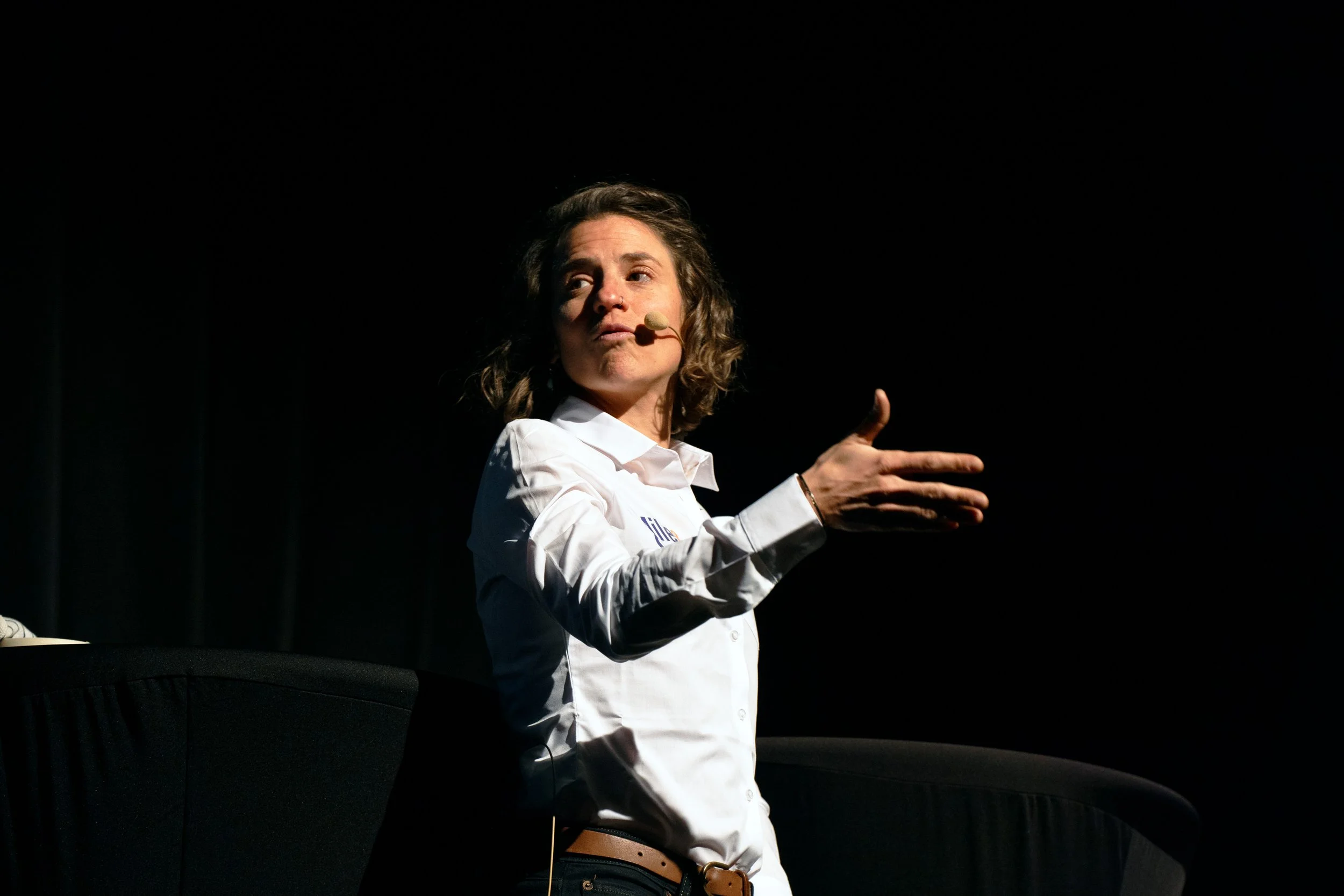 Woman speaking at a presentation, wearing a white shirt, in a dark light.