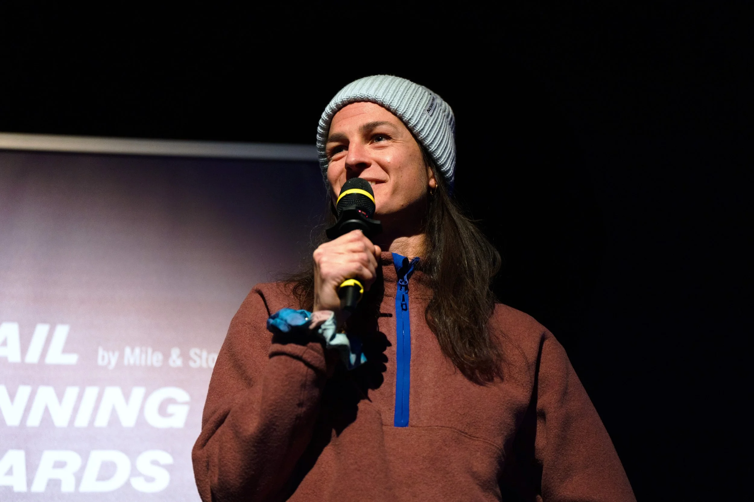 A woman speaking into a microphone at a ceremony or event, wearing a gray cap and brown sweatshirt, with a dark background and a screen behind with partially visible text.