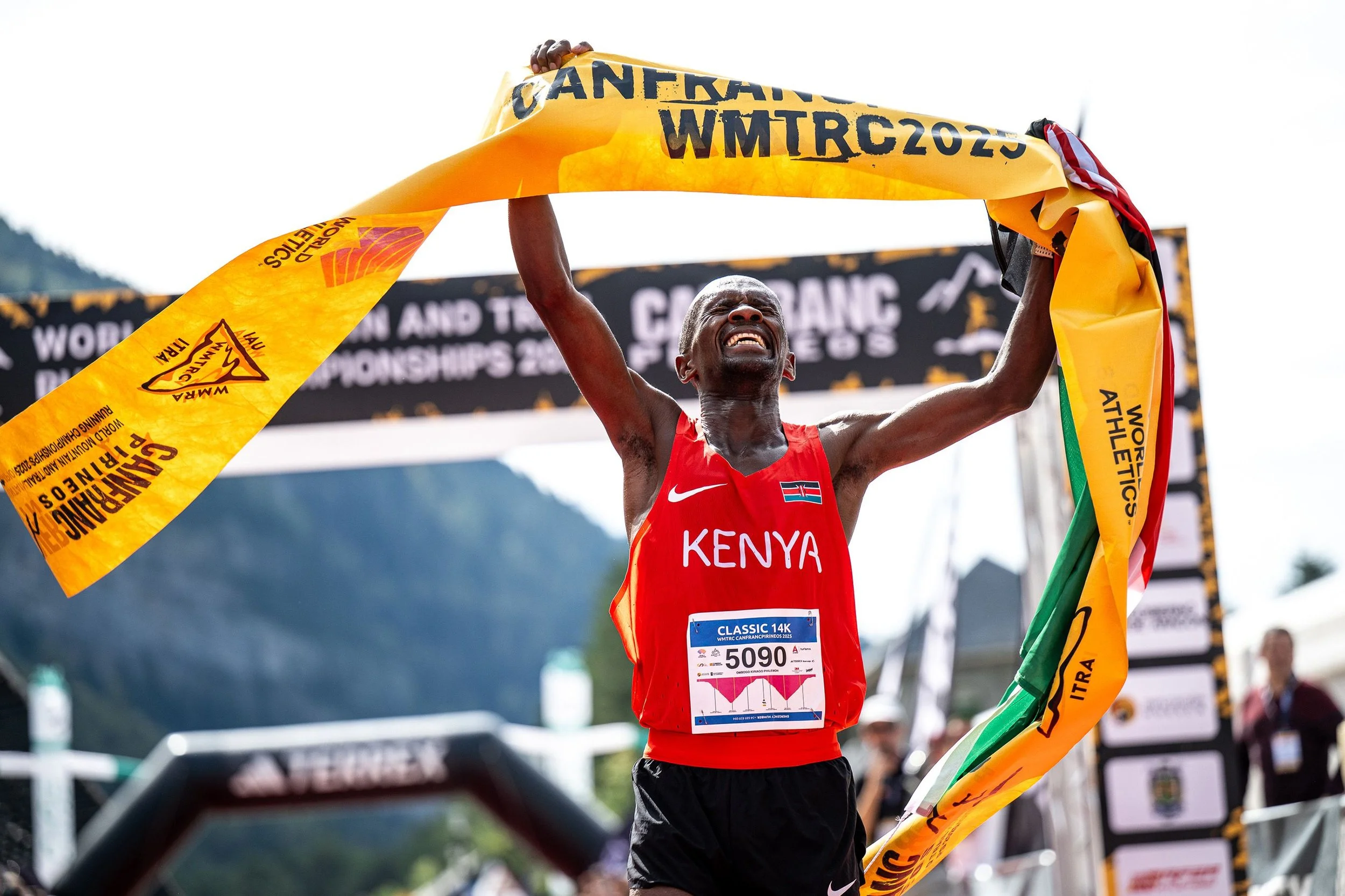 A Kenyan runner wins a 14 km race, raising the finish banner with an expression of victory, wearing a red jersey with "KENYA".