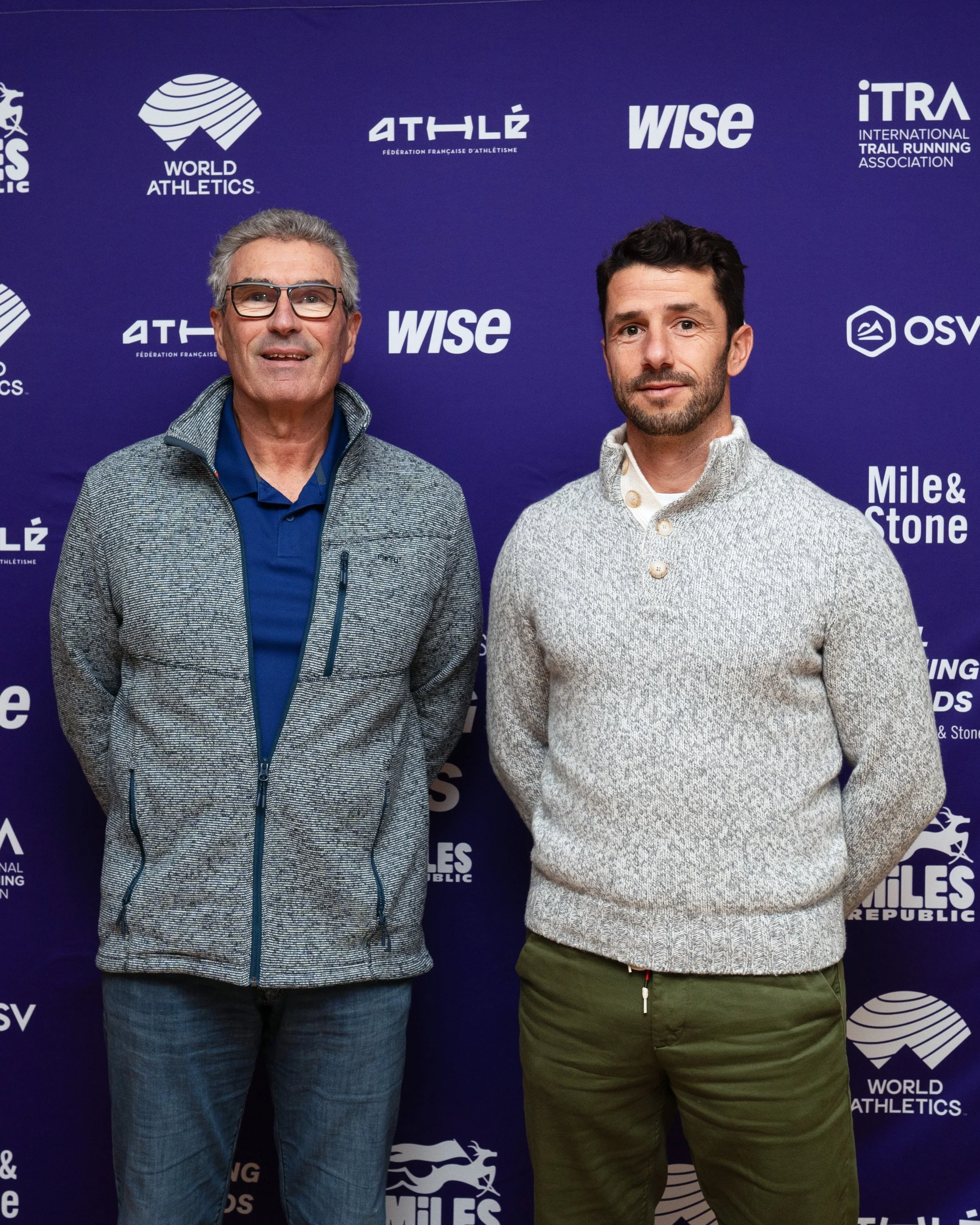 Two men posing for a photo in front of a blue background with logos related to athletics and trail running.