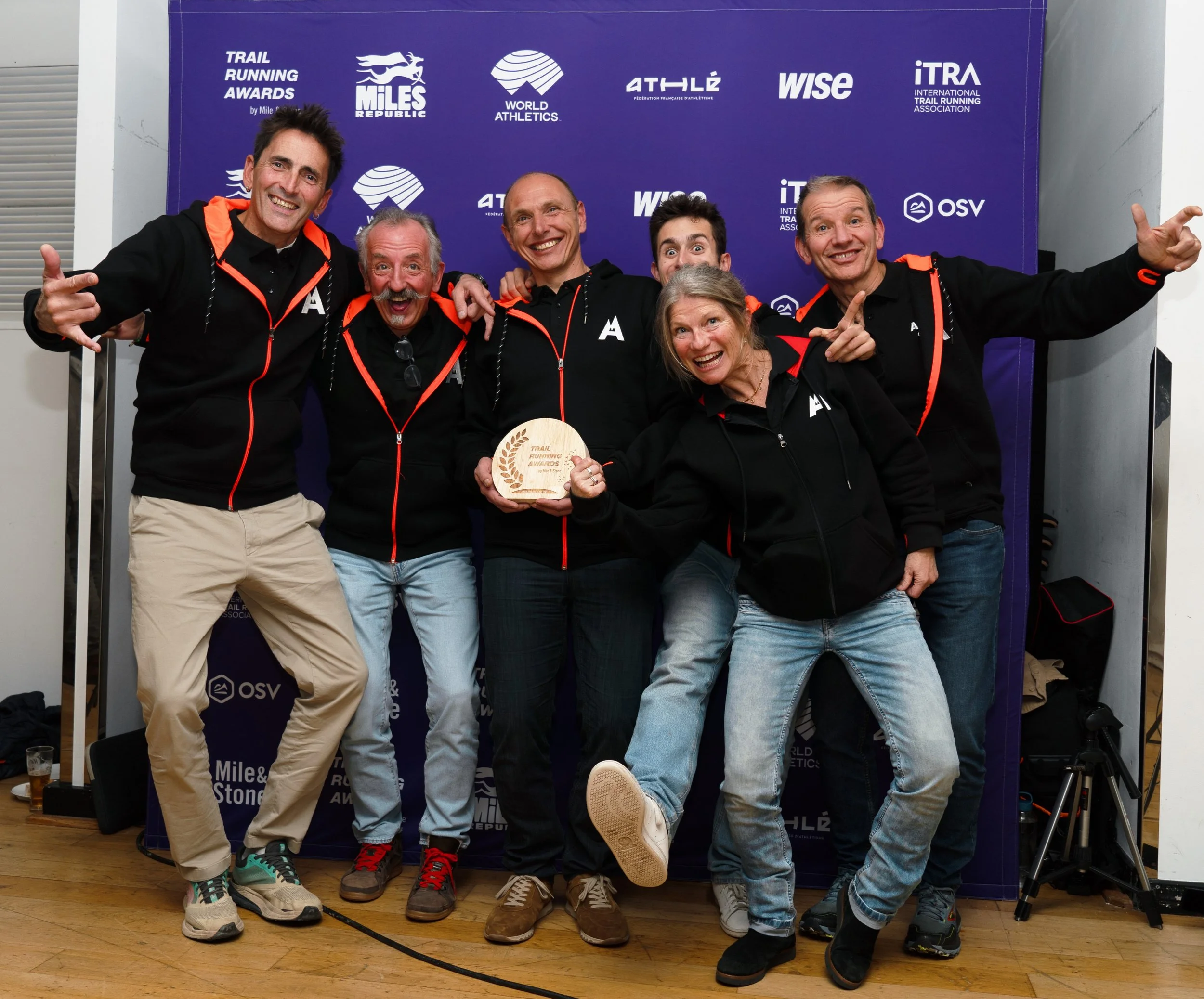 Group of six smiling people posing together at an awards ceremony, wearing black clothing with orange detailing, in the background a purple sign with logos related to trail running.