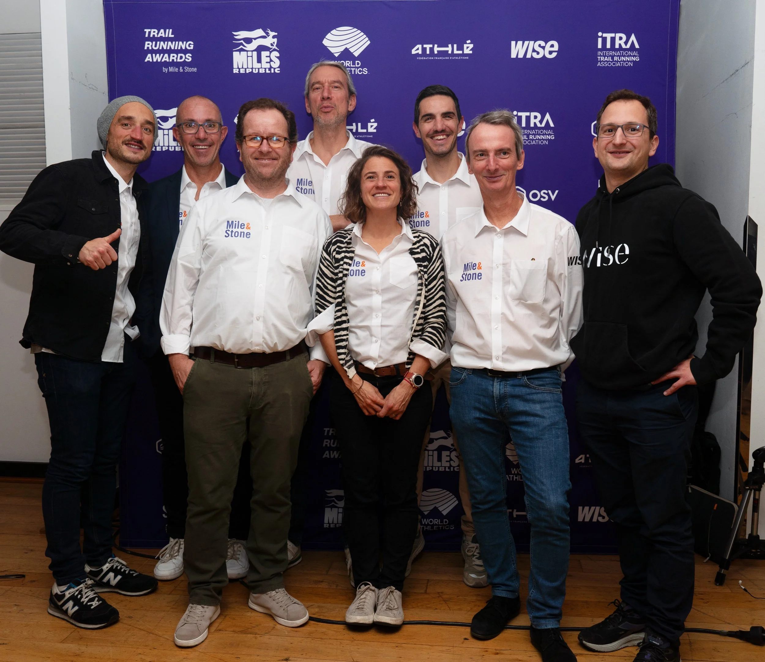 Group of nine posing for a photo at an awards ceremony, in front of a purple background with logos related to running and sporting events.
