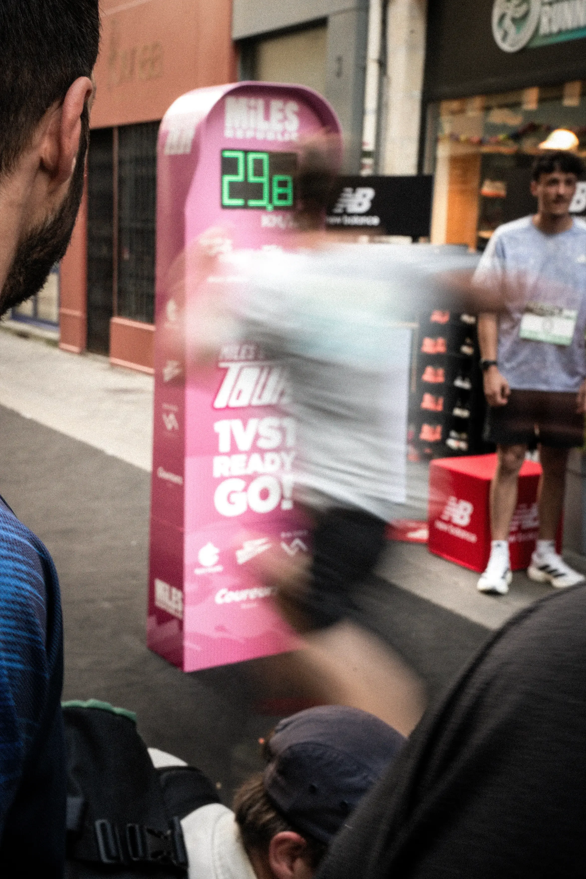 A group of people around a marathon stand. A man walks quickly past a water or refueling station, with a display showing 29 degrees Celsius. The station is decorated in pink with race logos. Runners are wearing sportswear and some have bibs.