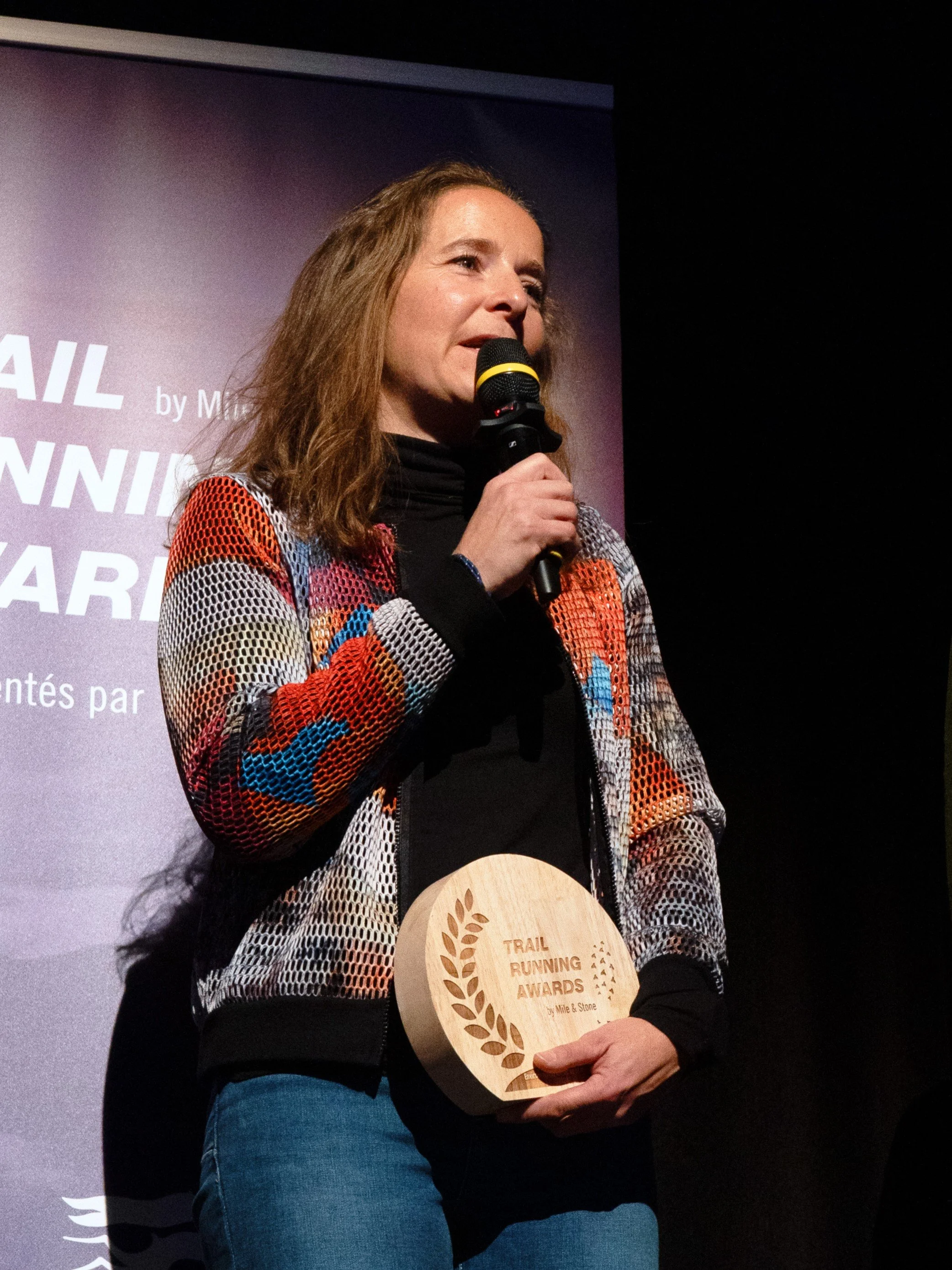 A woman holds a wooden trophy at a Trail Running Awards ceremony, speaking into a microphone.
