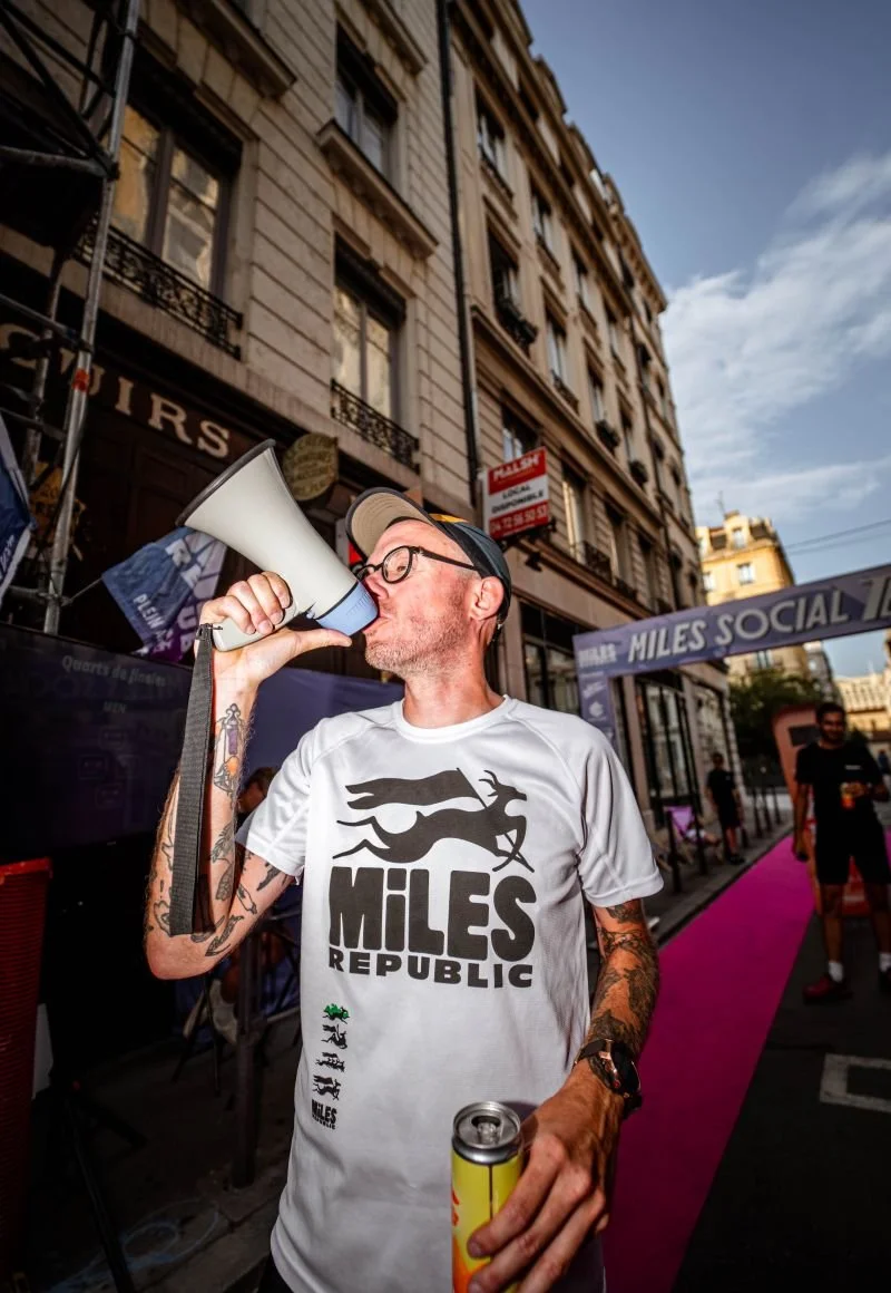 Man with tattoos, wearing a Miles Republic t-shirt and hat, speaking into a megaphone at an event in town.