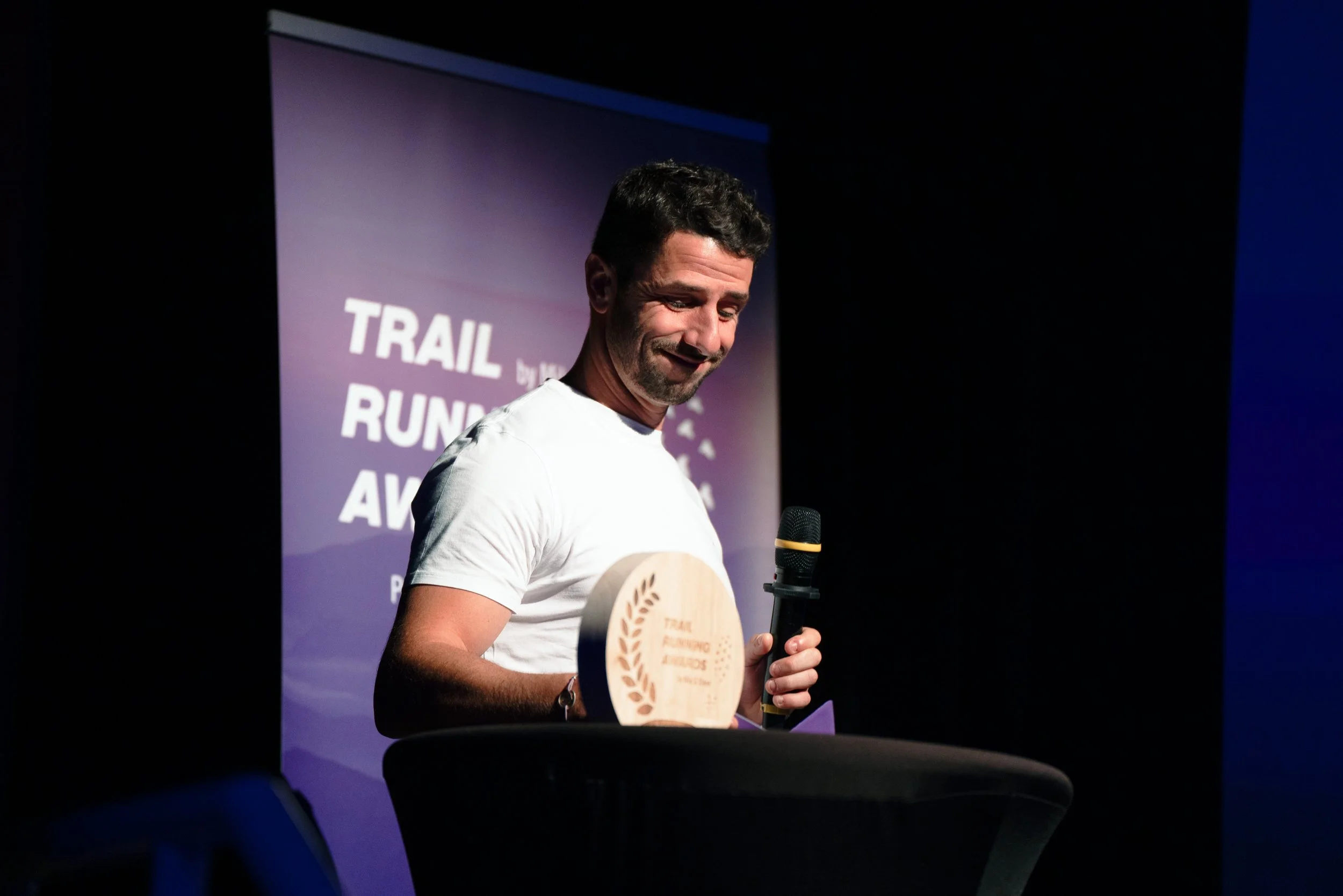 A smiling man at an awards ceremony, holding a wooden disc-shaped trophy inscribed "Trail Running Awards" and a microphone, with a sign in the background that reads "Trail Running Awards".