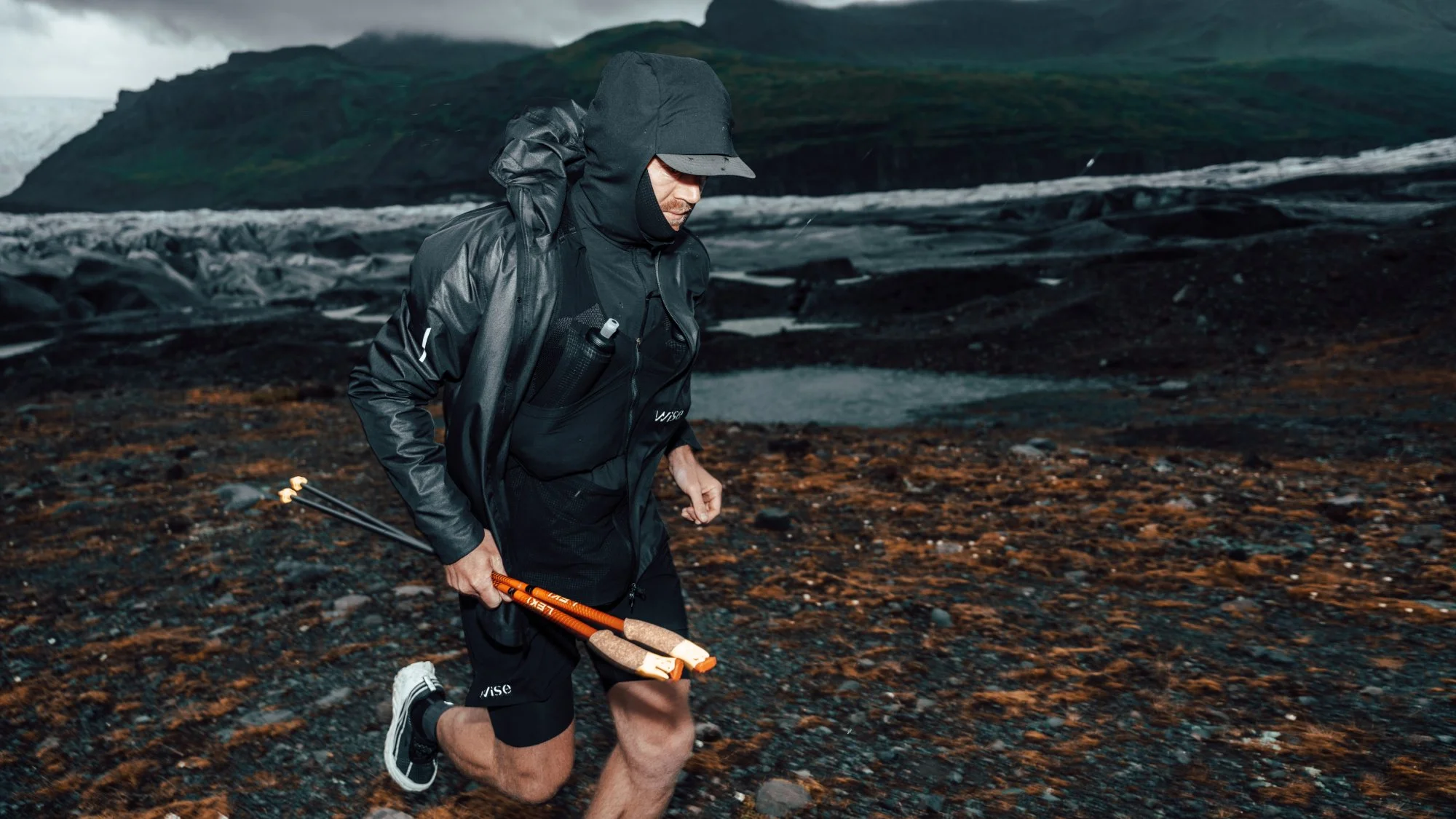 Man in rain gear running over rocky terrain with a mountainous landscape in the background.