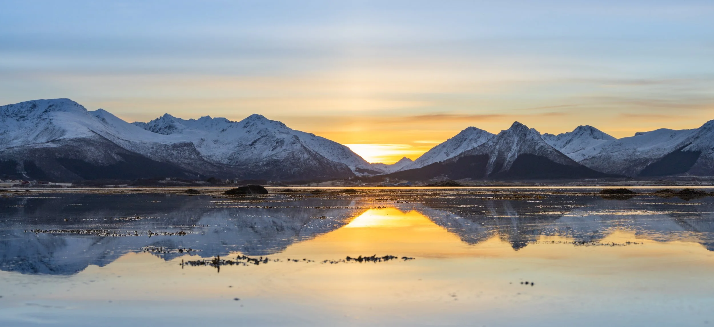 Snødekte fjell med refleksjon i stille vann under solnedgang.