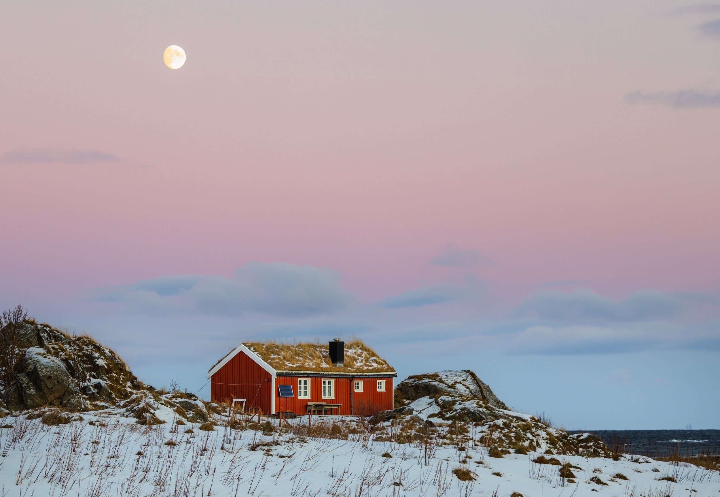 Rød hytte med snødekt tak på en øde kyst, omgitt av snø og steiner, under en lys måne og rosafarget himmel.