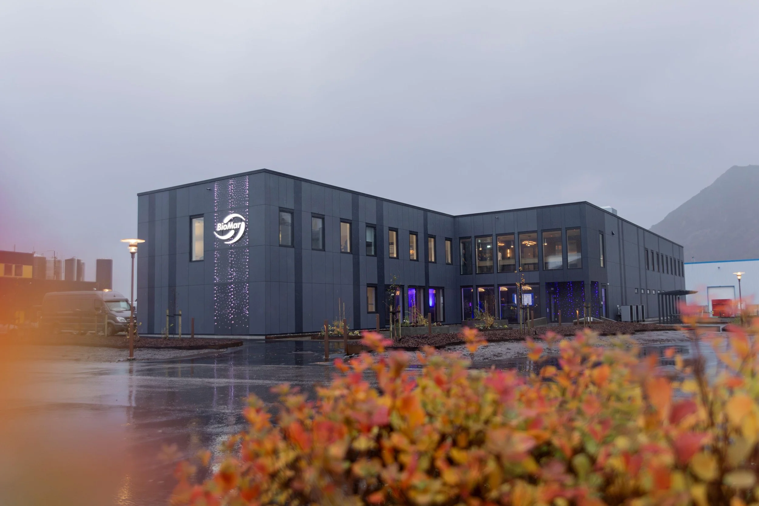 En moderne to-etasjers bygning med svart fasade og store vinduer, dekorert med blå og hvite lys; uteområdet er vått og omgitt av planter og gatelykter, med fjell i bakgrunnen.