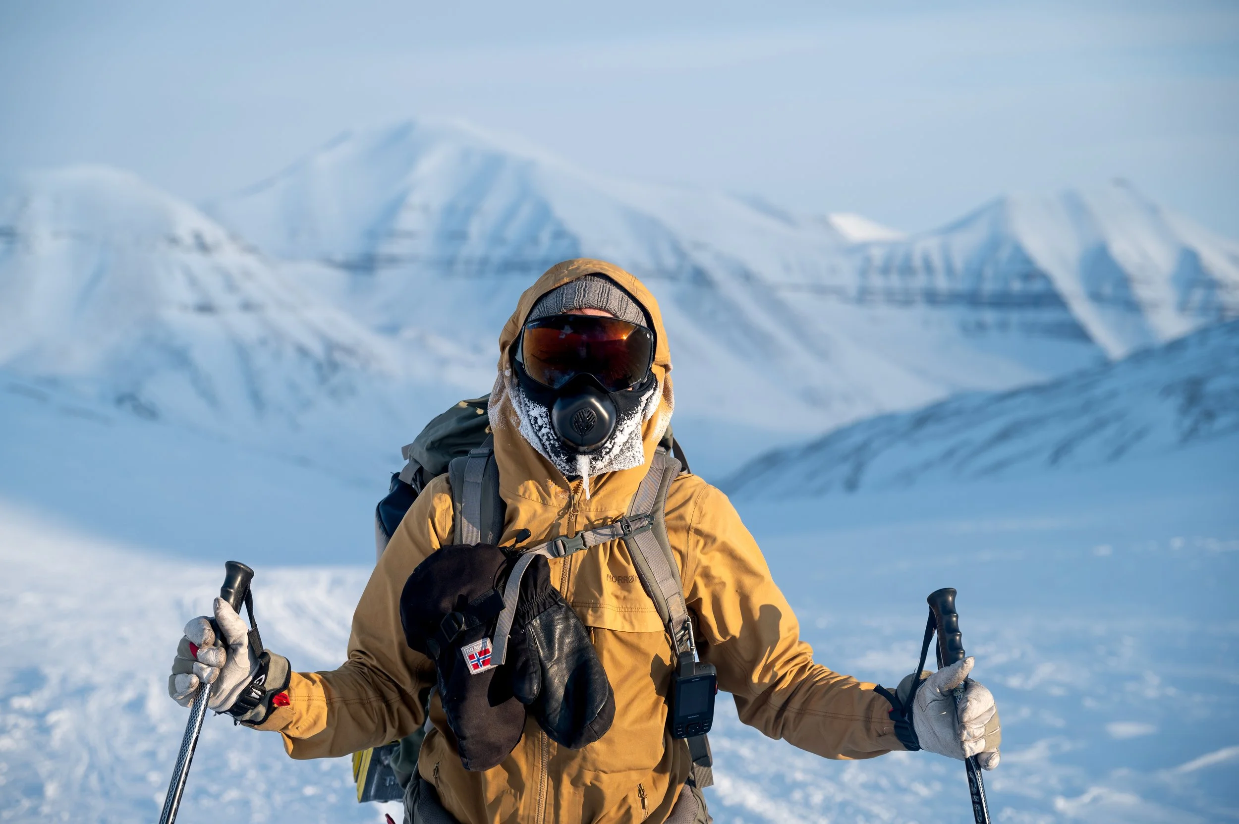 Anders Ottosen klædt i ekspeditionsudstyr med skibriller og åndedrætsværn, står i sneklædt landskab med snedækkede bjerge i baggrunden, og holder skistave.